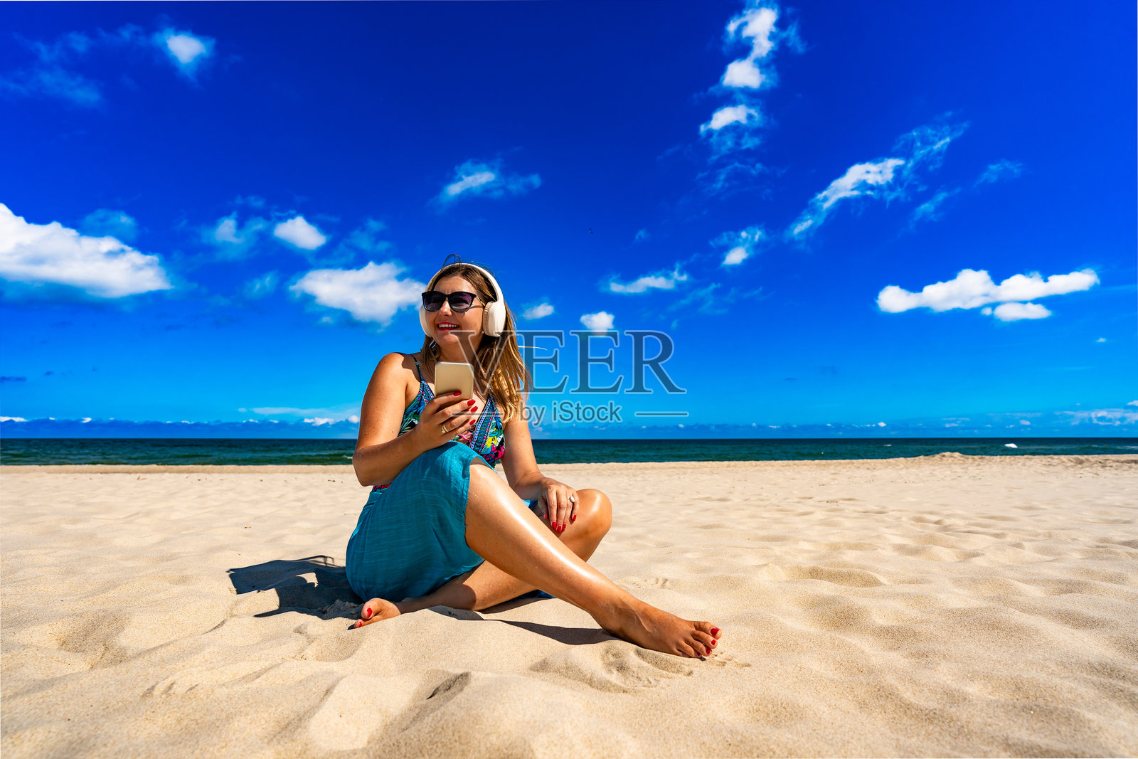 夏季音乐时光。美丽的成熟的女性坐在白色沙滩上，戴着耳机听音乐。照片摄影图片
