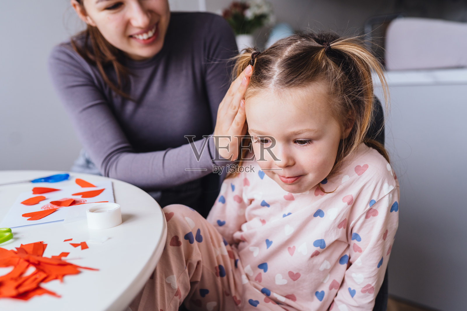 母亲温柔地抚摸女儿的头发，她们坐在装满手工艺品的桌子旁。小女孩专注于自己的艺术作品，感受到温暖的鼓励，这是一个充满关爱的家庭时刻。照片摄影图片
