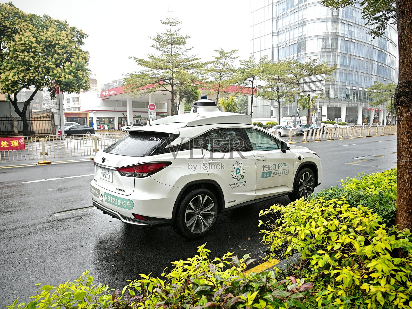 自动驾驶白色汽车带有车顶传感器，在雨天的湿滑城市街道上停靠，周围有绿植和建筑。照片摄影图片
