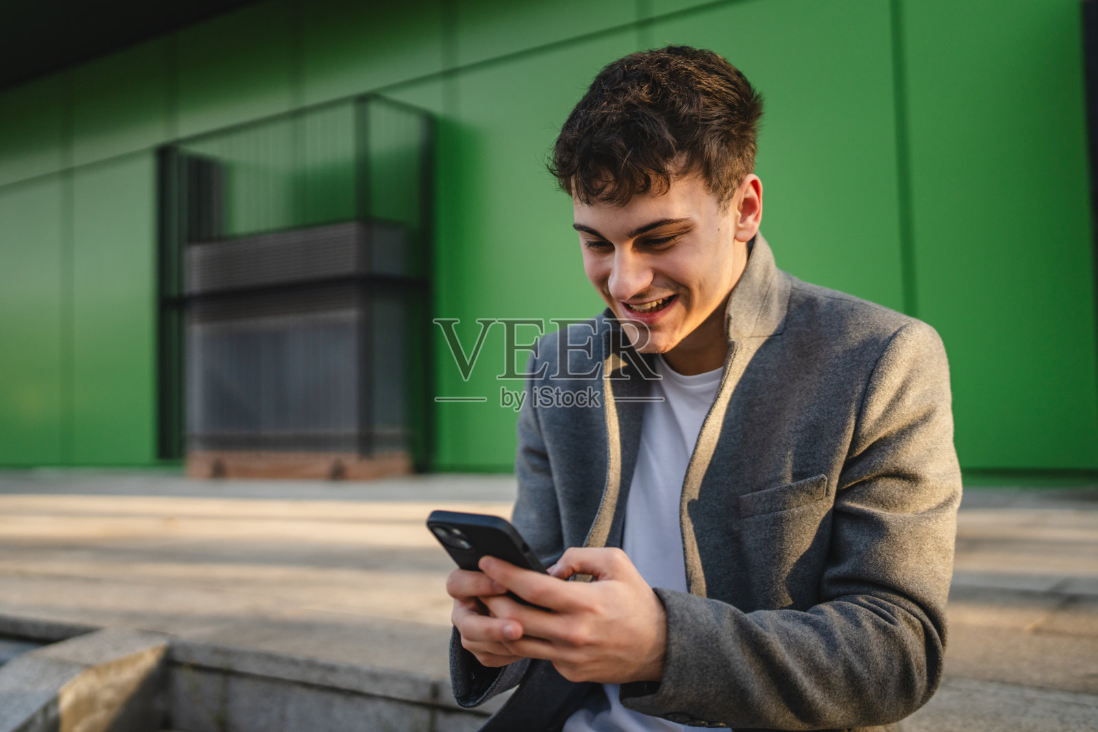 年轻快乐的男子坐在楼梯上使用手机，背景是绿色墙壁。照片摄影图片