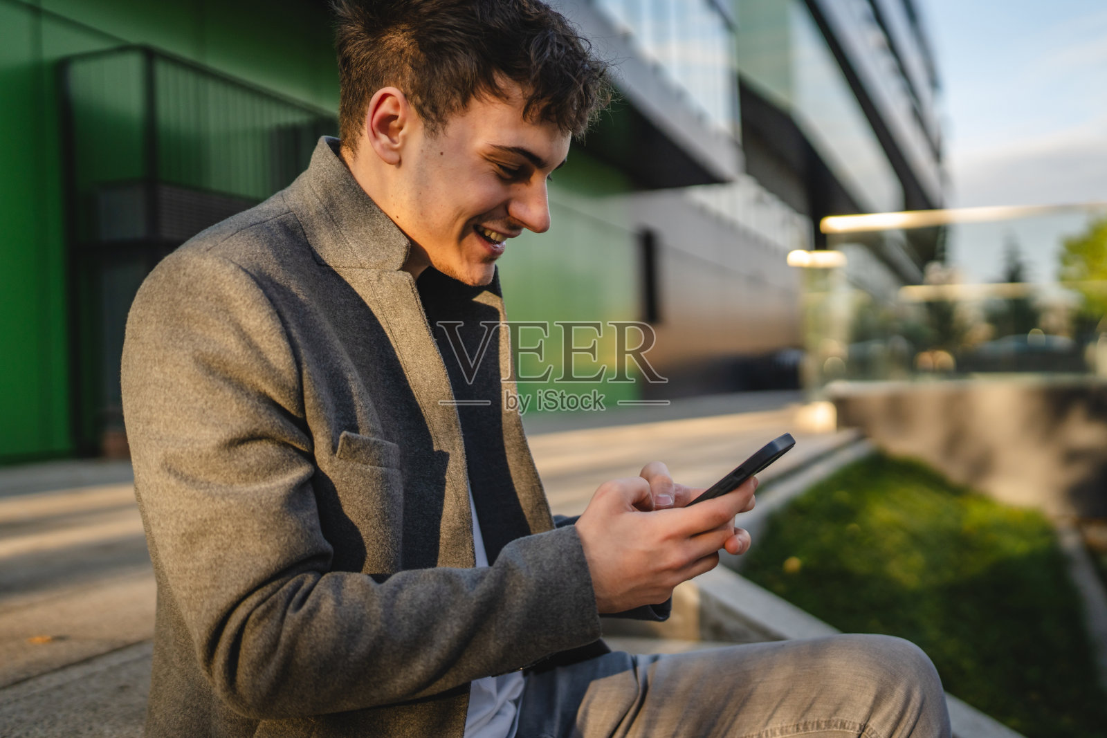 年轻快乐的男子坐在楼梯上使用手机，背景是绿色墙壁。照片摄影图片