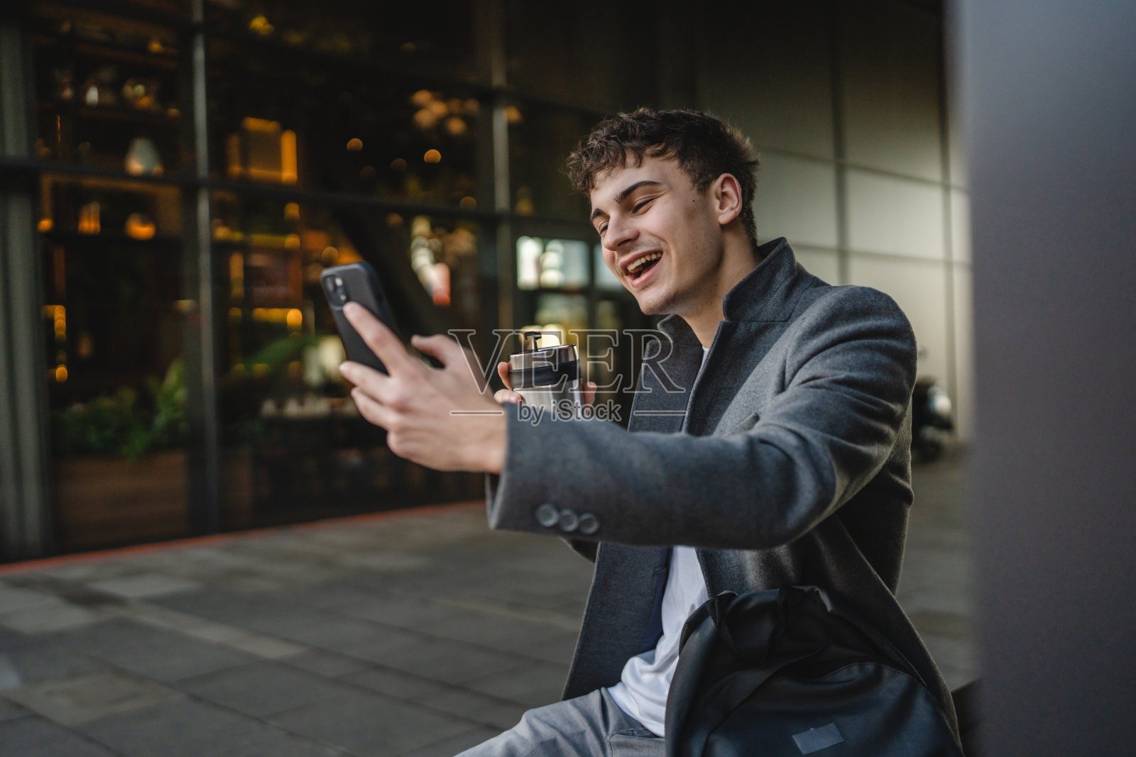 年轻男子坐在长椅上喝咖啡，正在用手机进行视频通话照片摄影图片