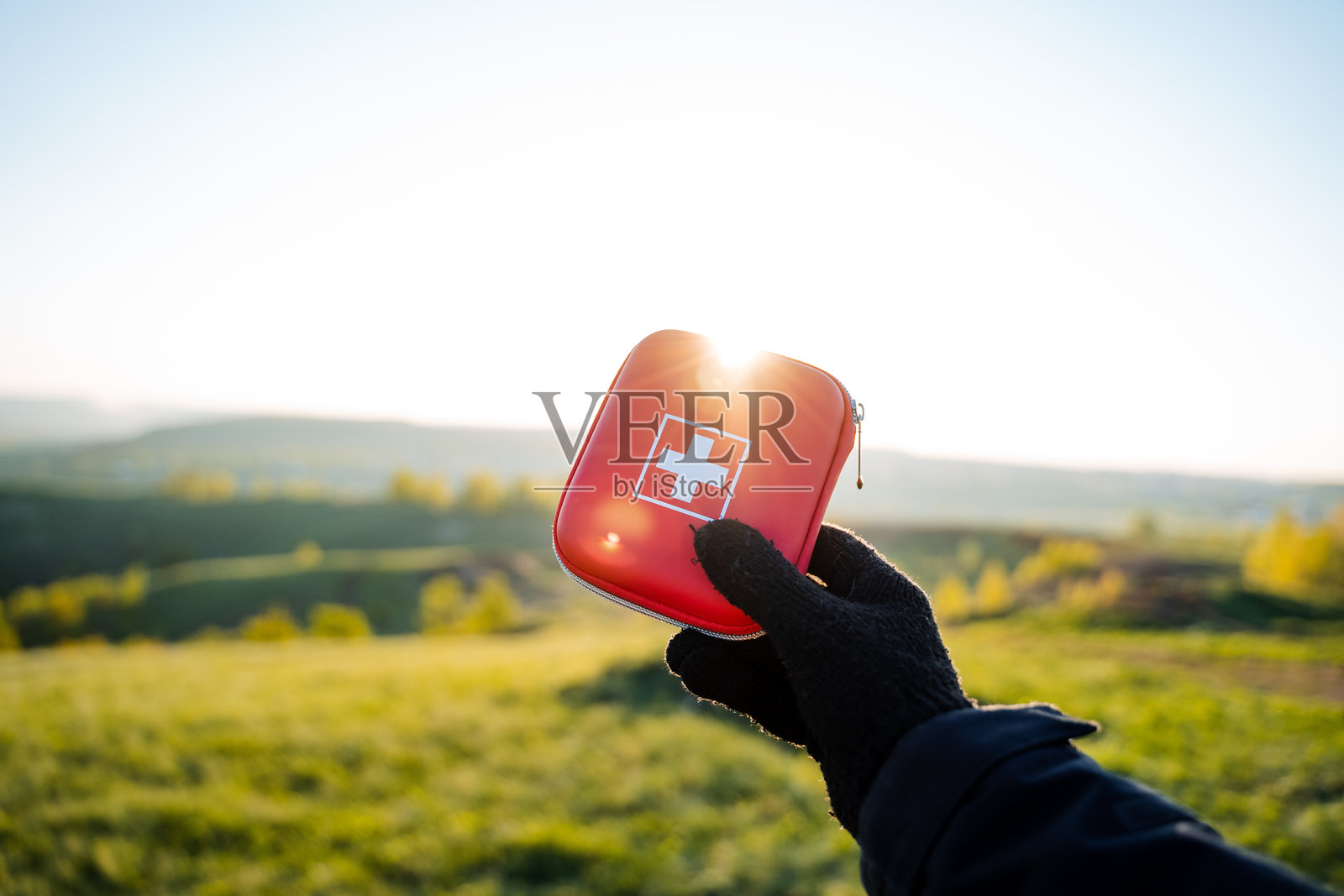 戴手套的手握着红色急救包，背景是户外自然风景，阳光照耀着旅行或户外活动中安全、健康和准备所需的重要物品。照片摄影图片