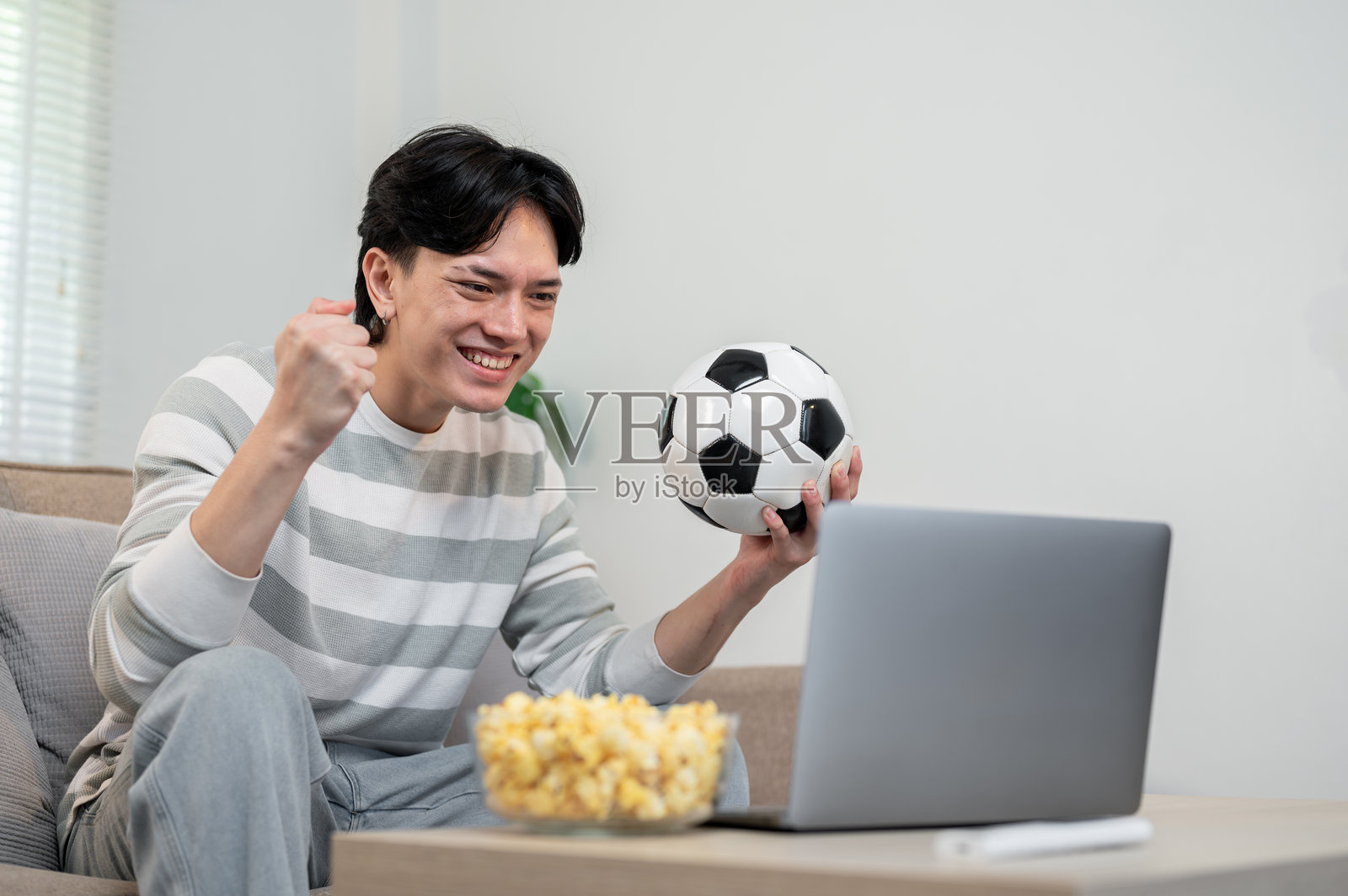 一位快乐的亚洲男子在家通过笔记本电脑观看他最喜欢的足球队的比赛。照片摄影图片