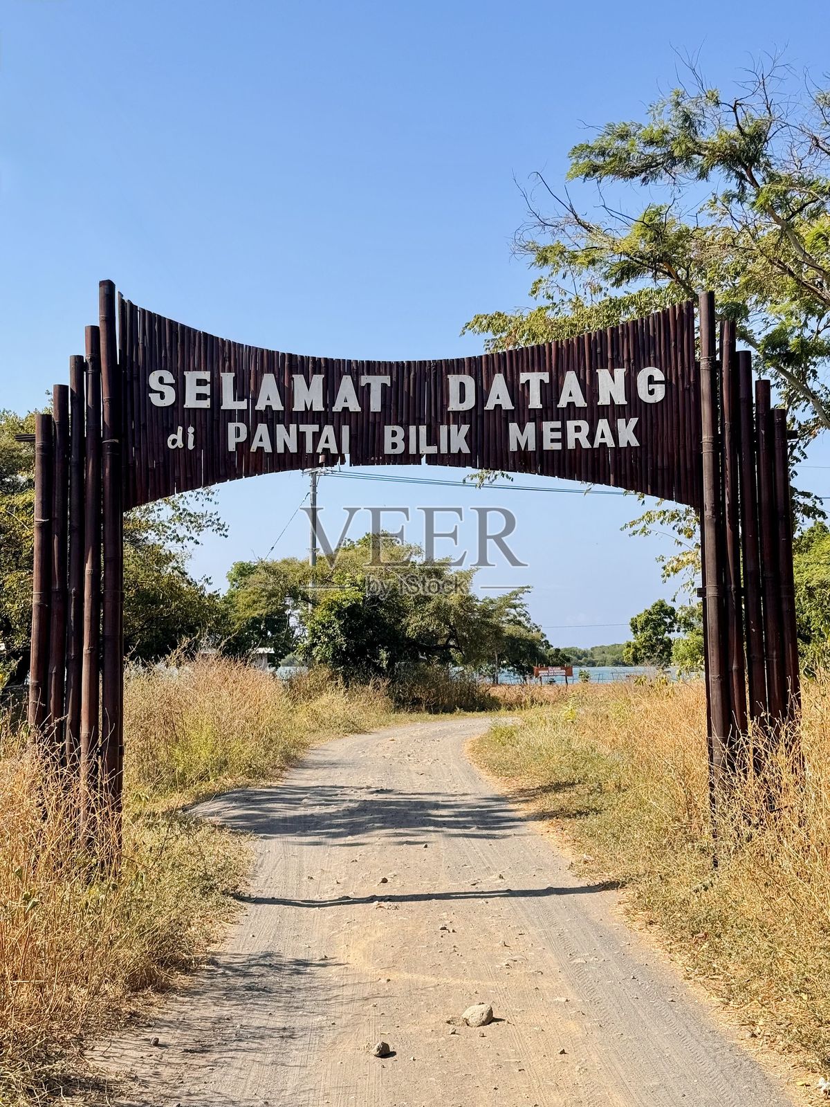在尘土飞扬的乡村道路上，巴厘克梅拉克海滩入口处的欢迎标志，周围是干枯的草地和树木。— 海岸旅游，木制拱门，热带目的地，印度尼西亚，户外旅行，夏季度假，风景如画的入口。照片摄影图片