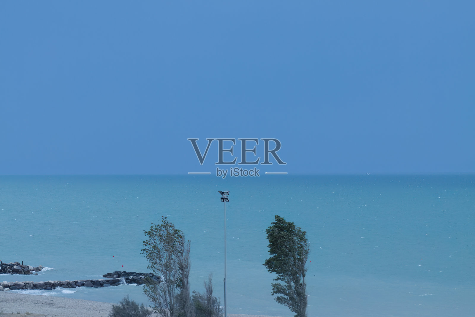 暴风雨中的蓝色海洋和蓝天，汹涌的海浪，沙滩上摇曳的树木，海天融为一体的蔚蓝色，暴风雨中的空旷沙滩，防波堤和高大的海浪，飞溅的浪花照片摄影图片