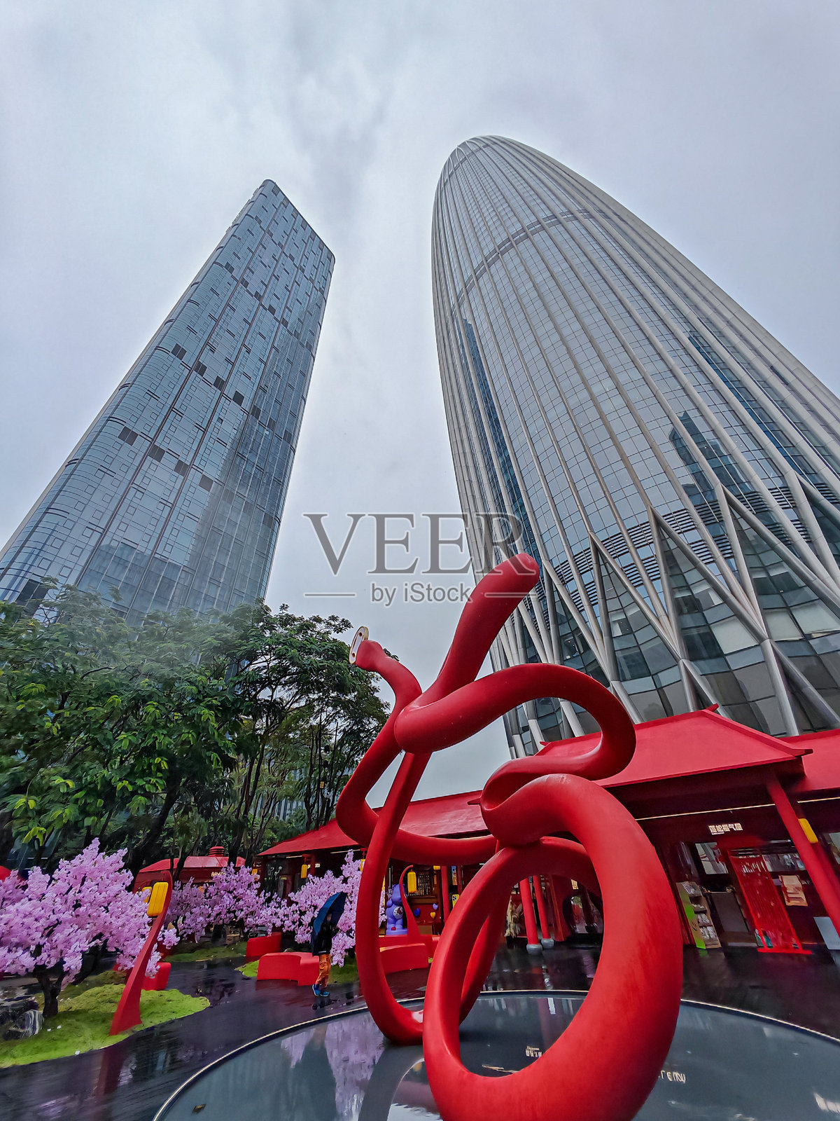 中国深圳城市景观，现代摩天大楼在阴云密布的天空下。前面是一个红色抽象雕塑和樱花，背景是传统的红色建筑。照片摄影图片