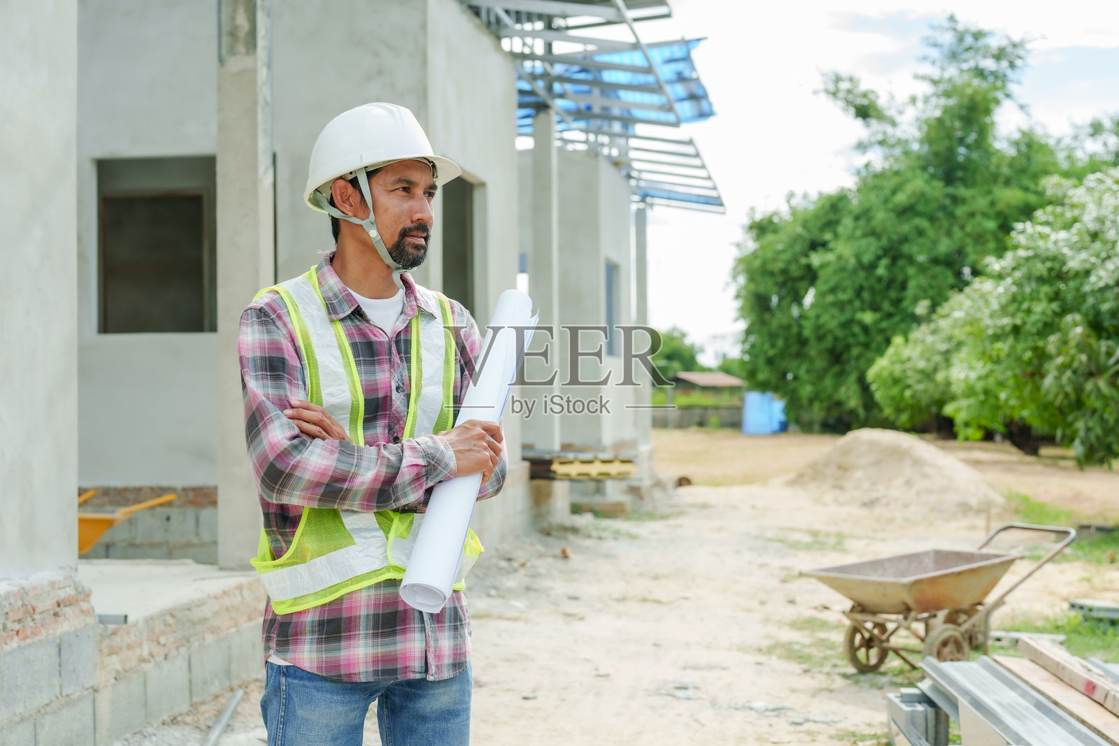 穿着安全头盔和反光背心的建筑师双臂交叉，手持蓝图，站在住宅建筑工地上。周围有未完工的房屋和建筑材料及设备。照片摄影图片