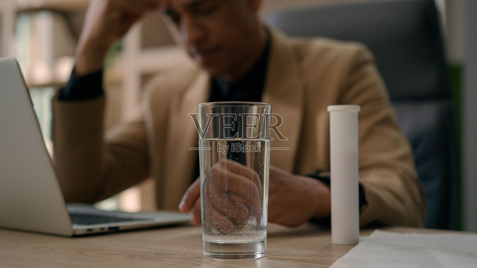 过度劳累的非裔美国商人将止痛药片扔进水杯中，药片在饮料中起泡，办公室桌上，痛苦的商人因头痛和宿醉而准备解药。照片摄影图片