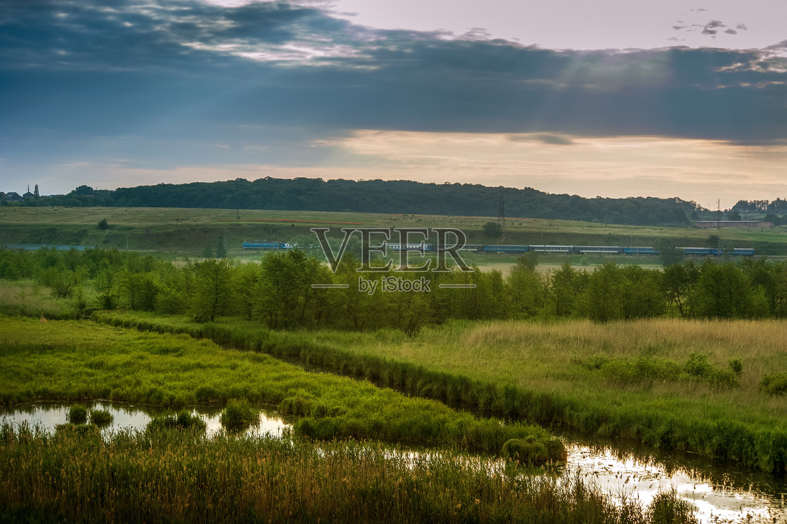 湿地、河流泛滥平原、广阔的绿色草地，远处的火车在阳光透过云层的柔和光线下，营造出宁静的氛围照片摄影图片