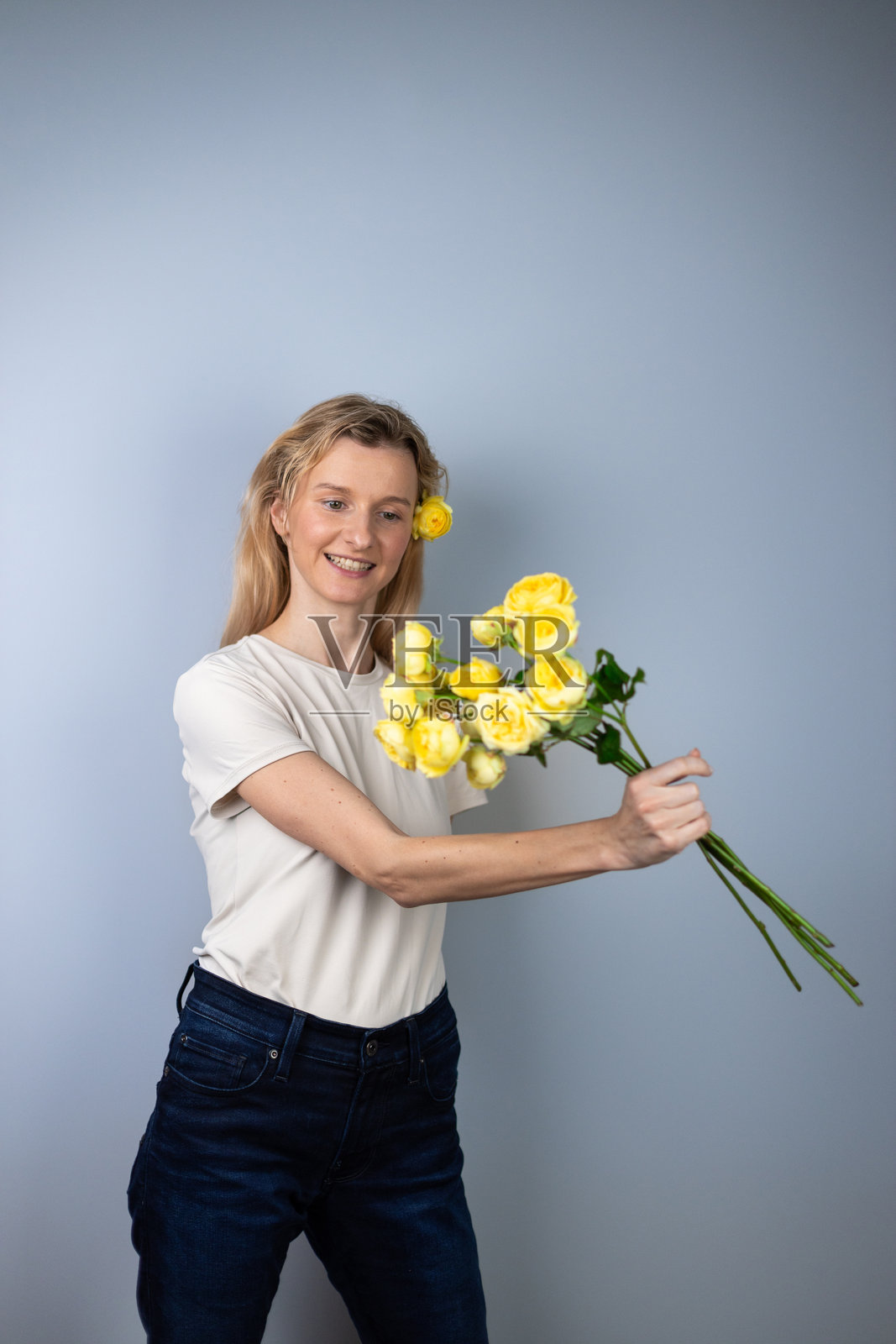 在浅灰色背景下，一位年轻的金发女孩穿着浅色T恤，面带微笑，向前抛洒着牡丹玫瑰和三月八日的黄色花朵。照片摄影图片