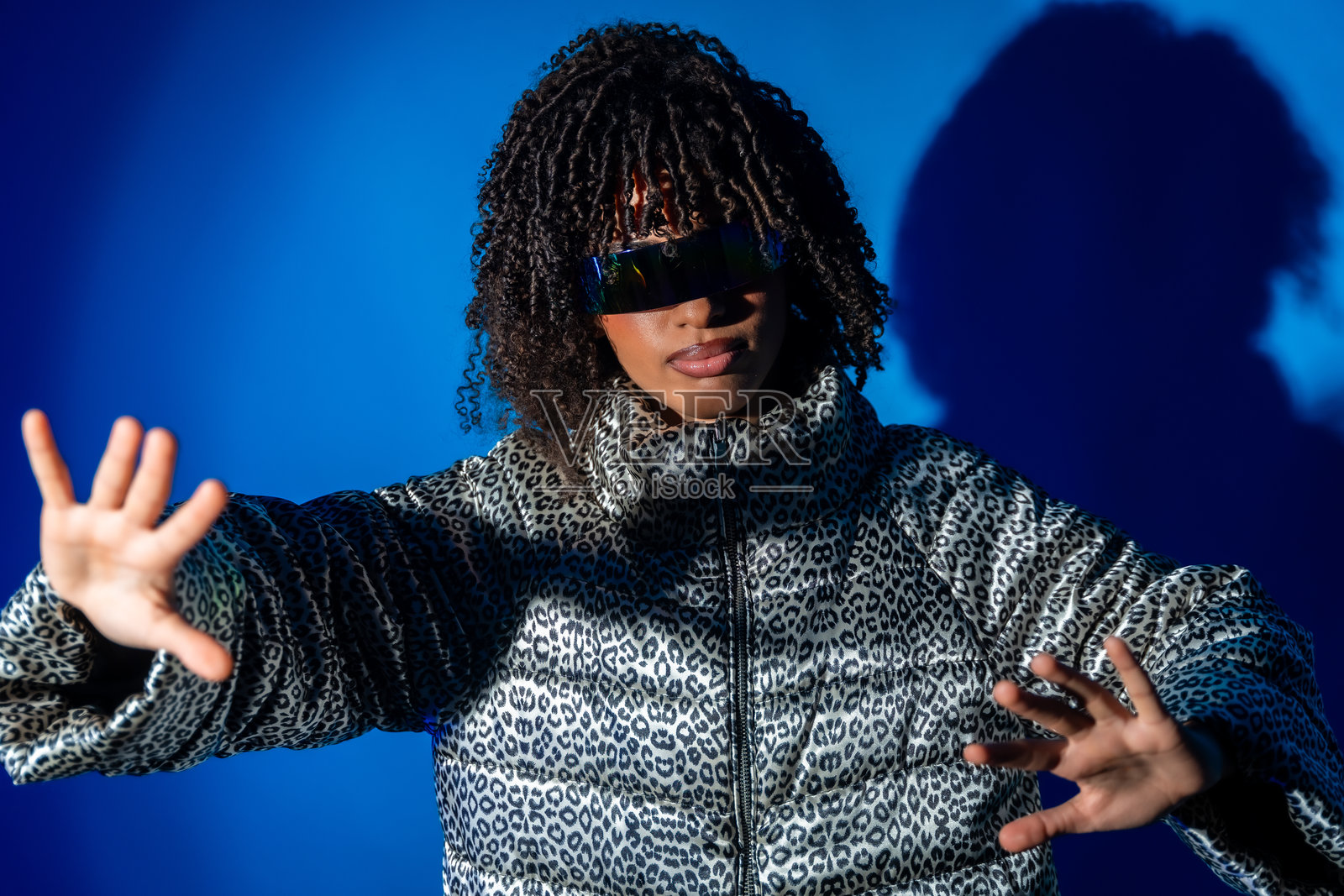 年轻时尚女性穿着豹纹夹克和未来感太阳镜，摆姿势于蓝色背景前照片摄影图片