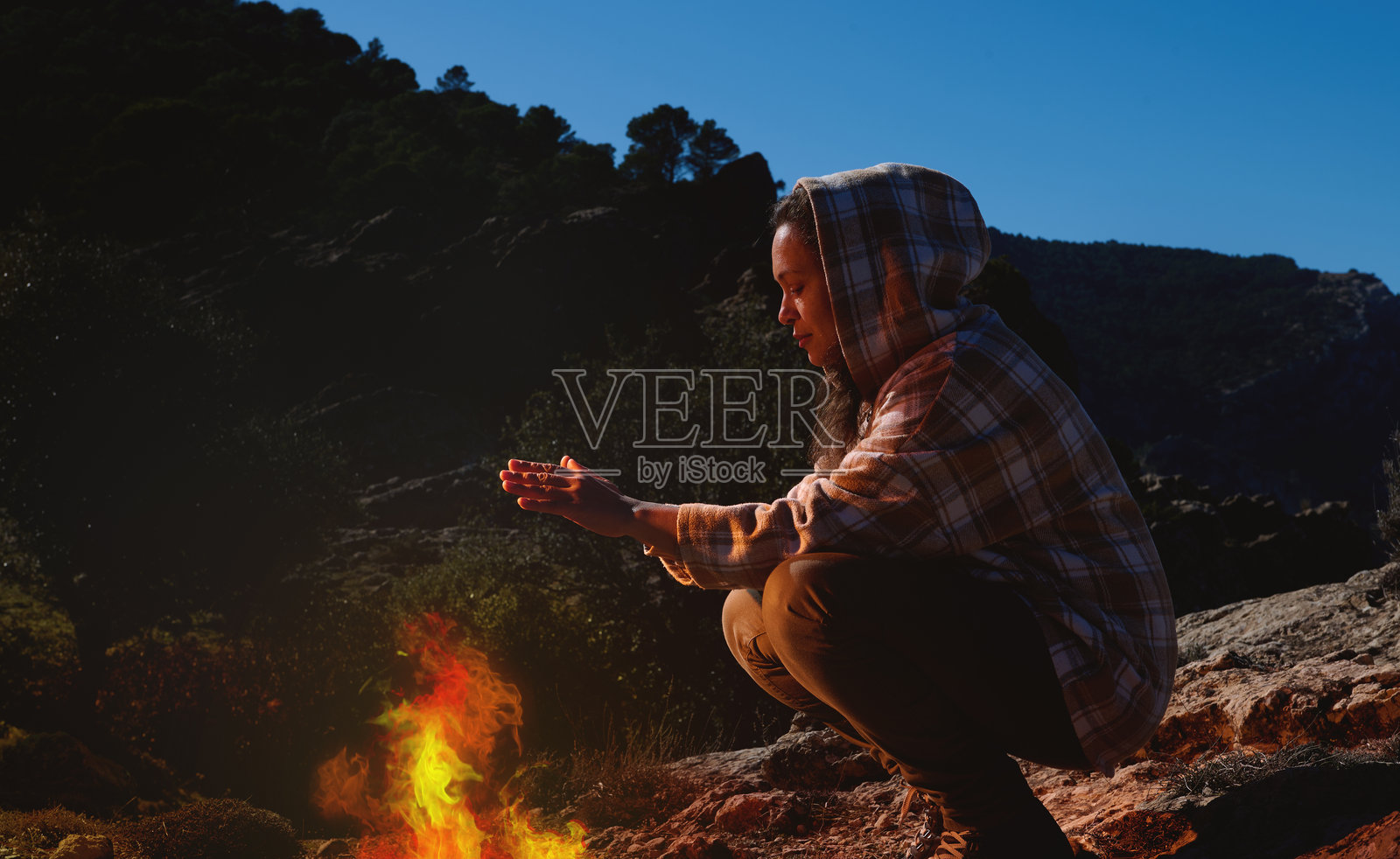 一个人穿着带帽夹克，坐在野外的篝火旁，享受傍晚时光。照片摄影图片