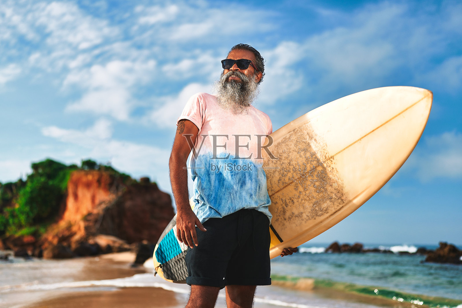 阳光明媚的海滩上，一位老年黑人男性手持冲浪板站立。戴着太阳镜的老年男性在海浪旁享受冲浪。积极的生活方式，老年人的冒险活动。照片摄影图片