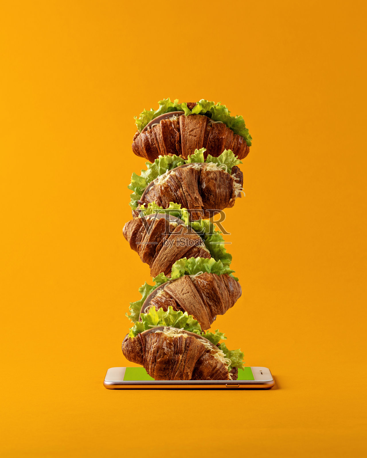 填充可颂和生菜堆叠成一座塔，从智能手机屏幕中冒出，背景为橙色。在线订餐和外卖的概念。照片摄影图片