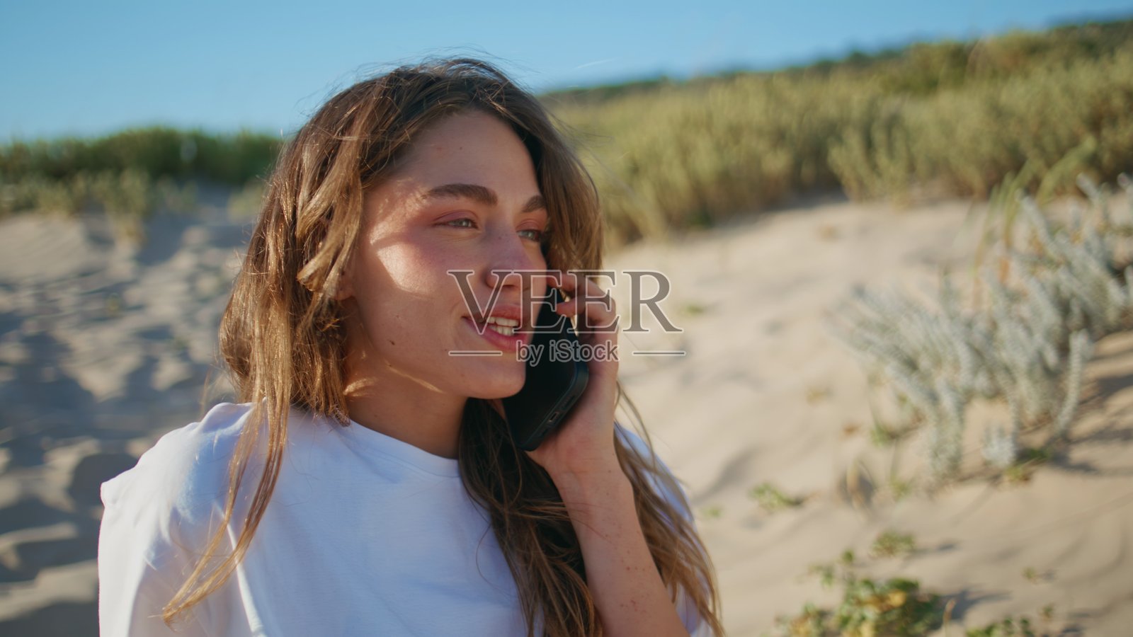 阳光明媚的女子在狂野的海滩上打电话特写。快乐的女孩在使用智能手机交谈。照片摄影图片