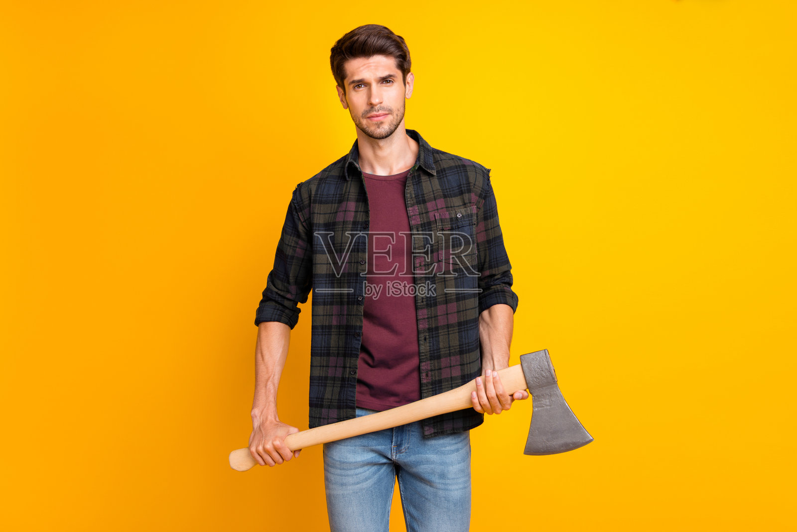 一个严肃的男子面无表情，准备使用他强壮的手臂握着的大斧，穿着休闲的格子衬衫，背景为黄色。照片摄影图片