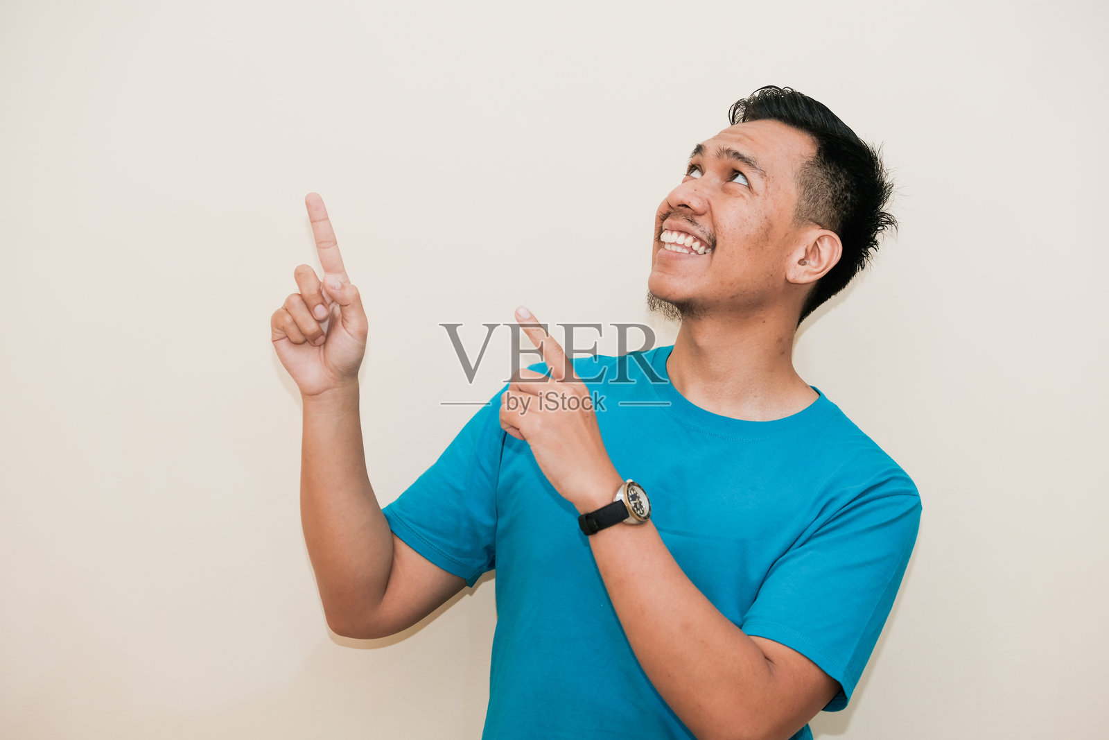一位穿着青绿色T恤的东南亚男子微笑着，右手用两个手指指向上方，背景为白色。照片摄影图片