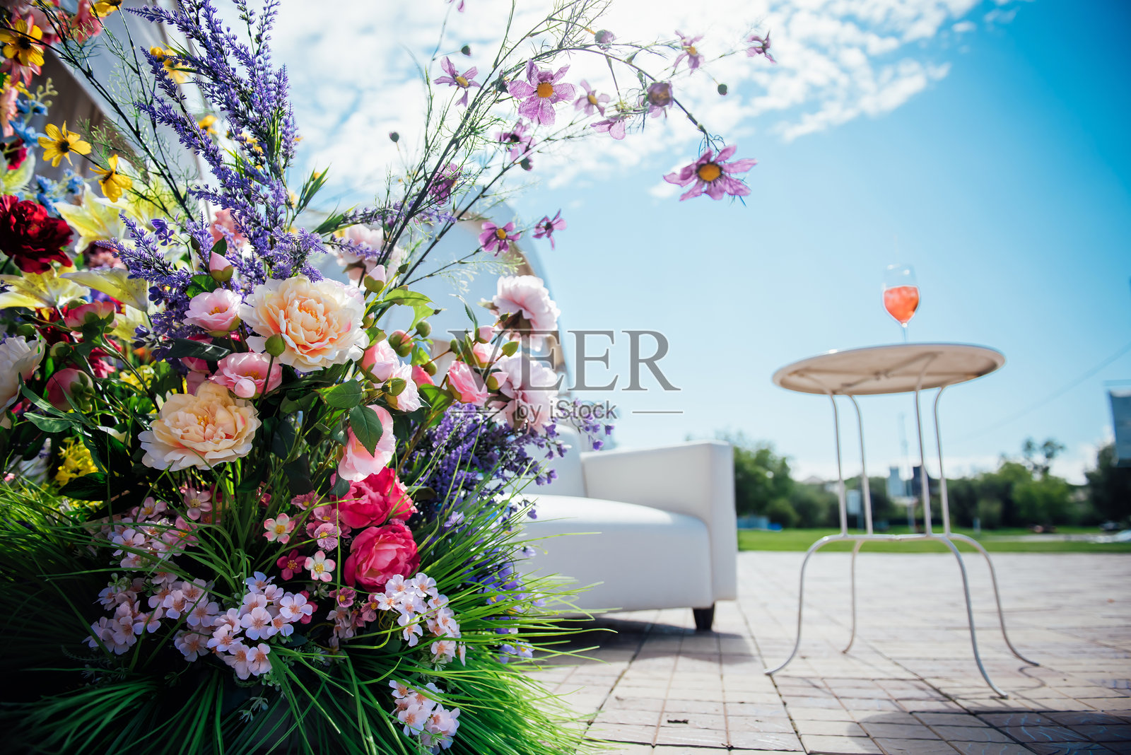 一个巨大的户外新鲜花卉花束装饰着一家高档餐厅的户外休闲区。一个舒适的户外座位区，周围有花朵和一张摆放着酒杯的桌子，蓝天映衬着这一切。照片摄影图片