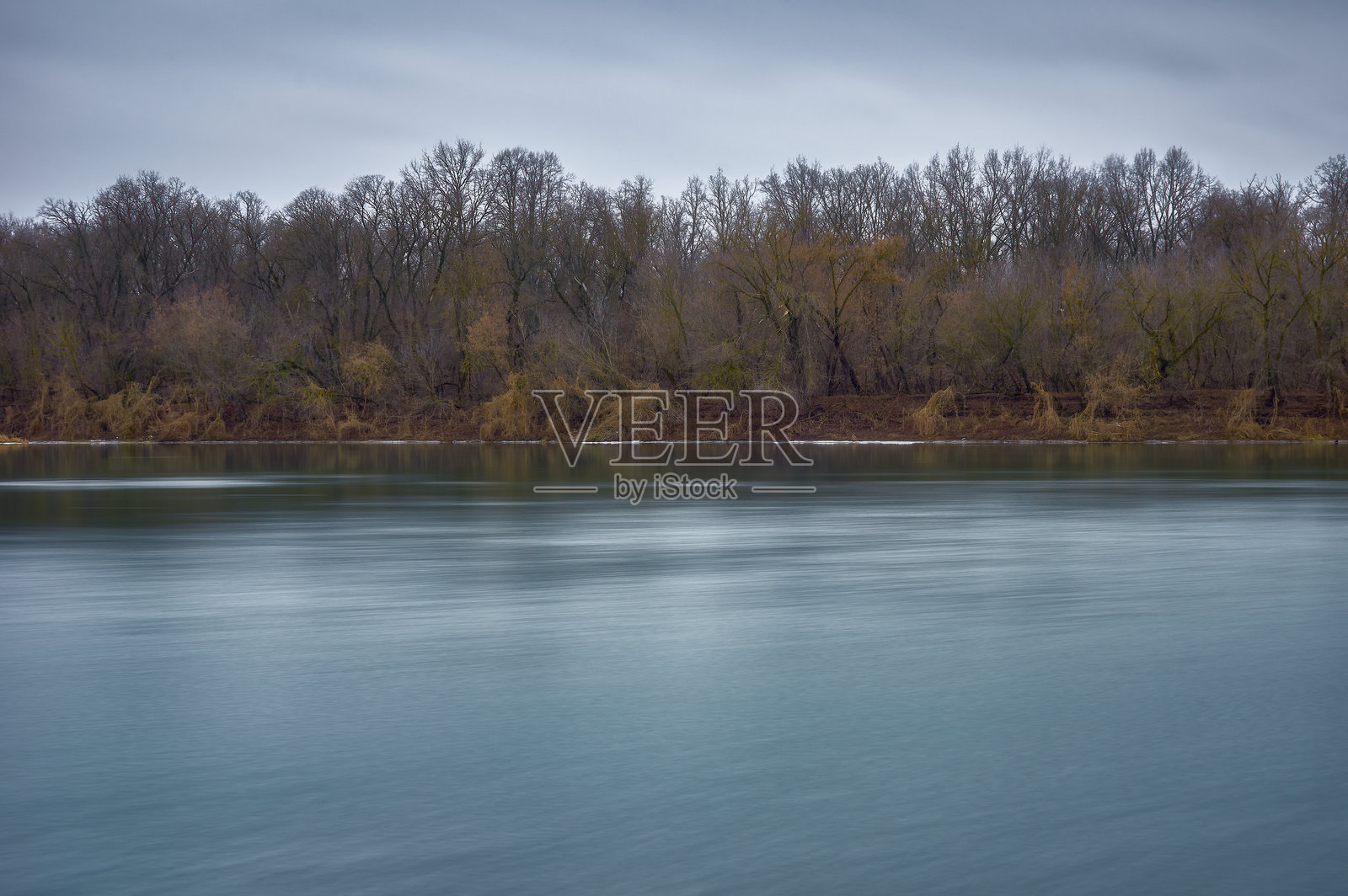 冬天，天气温暖，雪已经融化，河水缓缓流动，河岸上长满了草、灌木和树木，草已经枯萎，树木光秃秃的，天空阴云密布。照片摄影图片