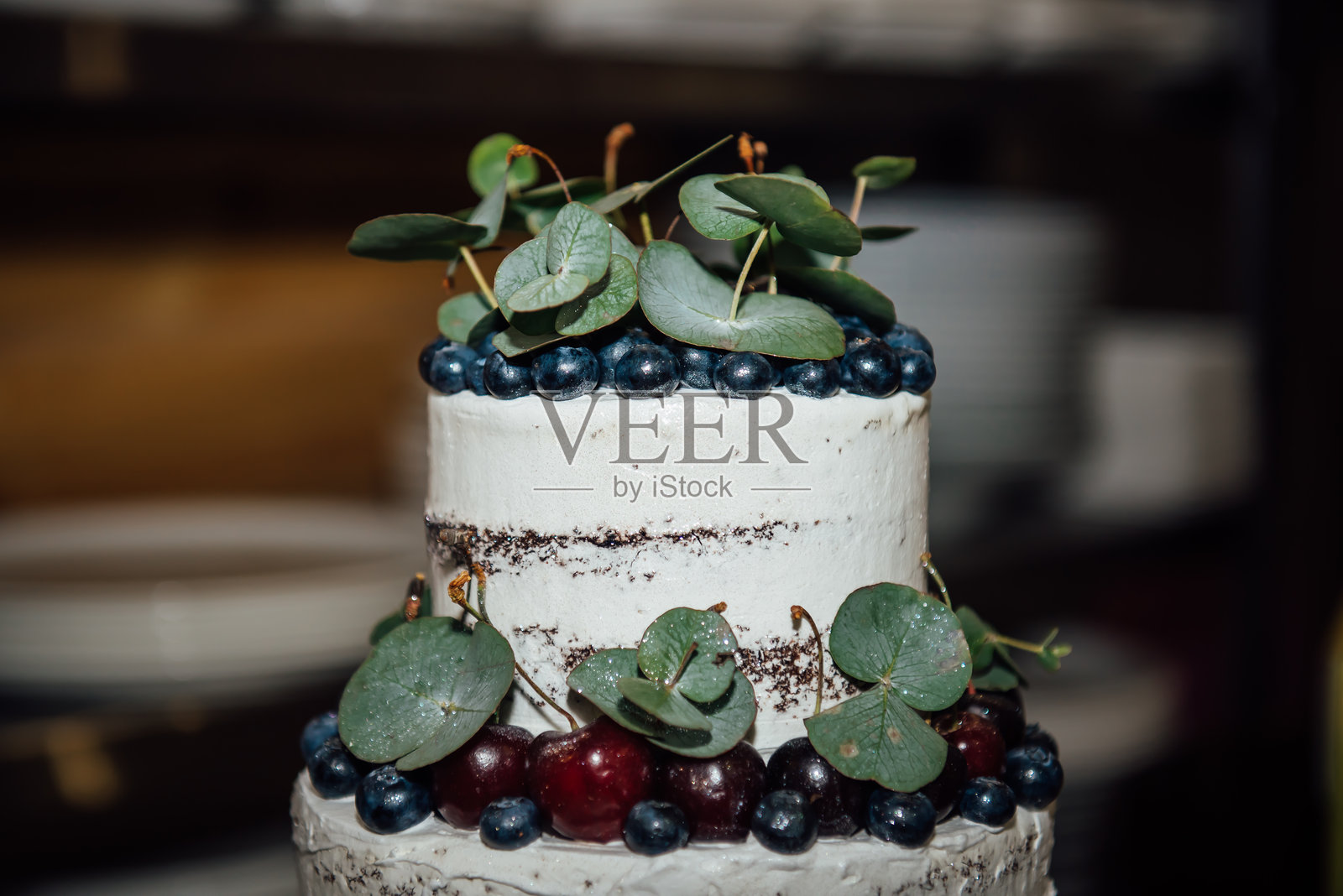 美丽的白色婚礼蛋糕，分为两层，装饰着新鲜的蓝莓、樱桃和绿叶。一个大型优雅的婚礼蛋糕的顶部。照片摄影图片