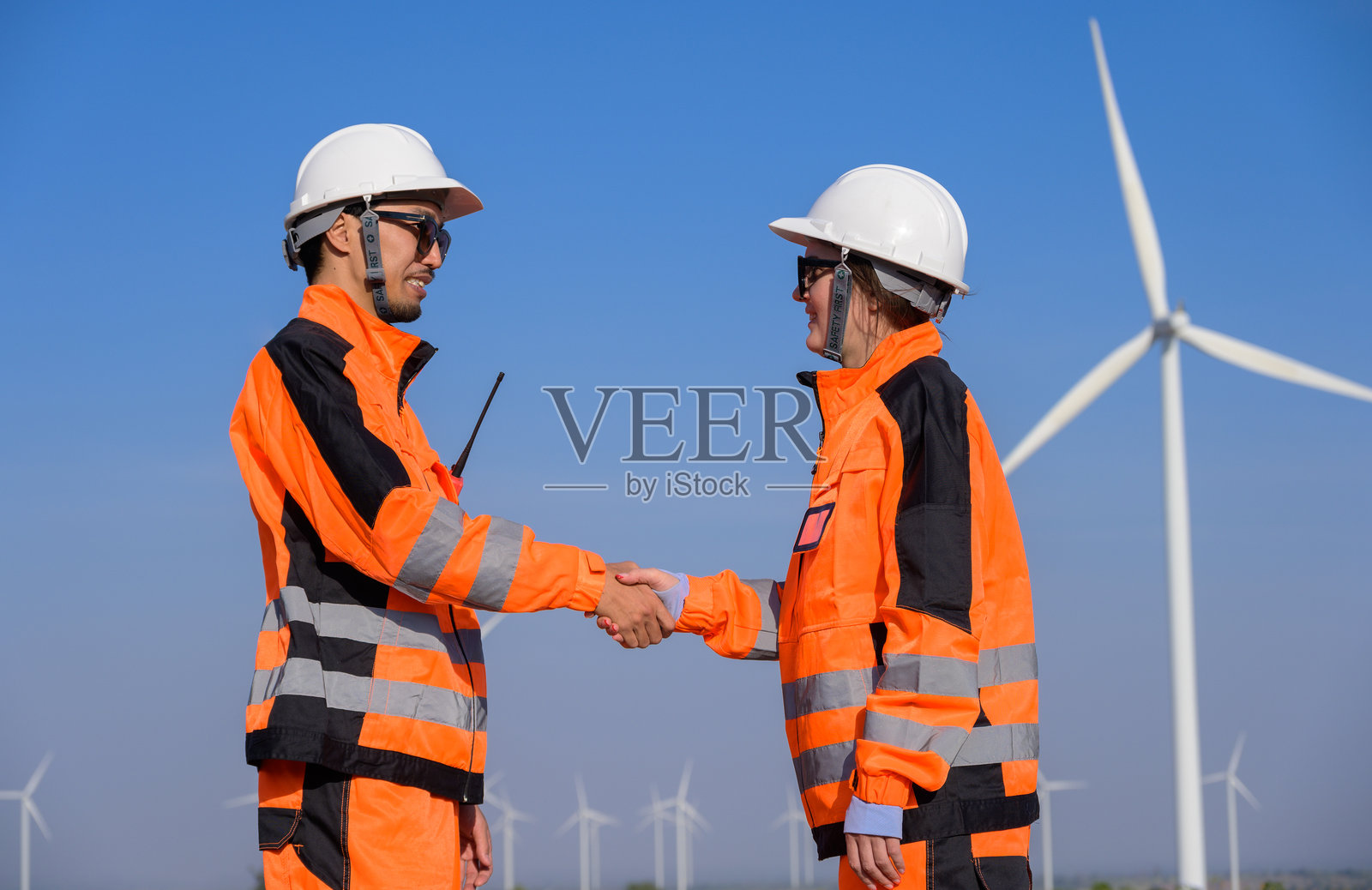 两名穿着橙色制服的工人在风力发电场握手。照片摄影图片