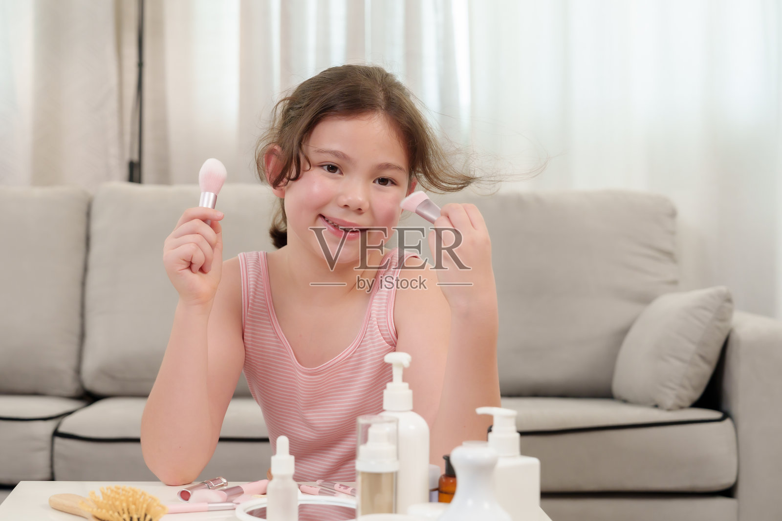 白人女孩学习化妆。一位快乐的年轻女孩在梳妆台前玩耍，手里拿着两支化妆刷，周围摆满了美容产品。这个瞬间捕捉了她对美的快乐和好奇。照片摄影图片