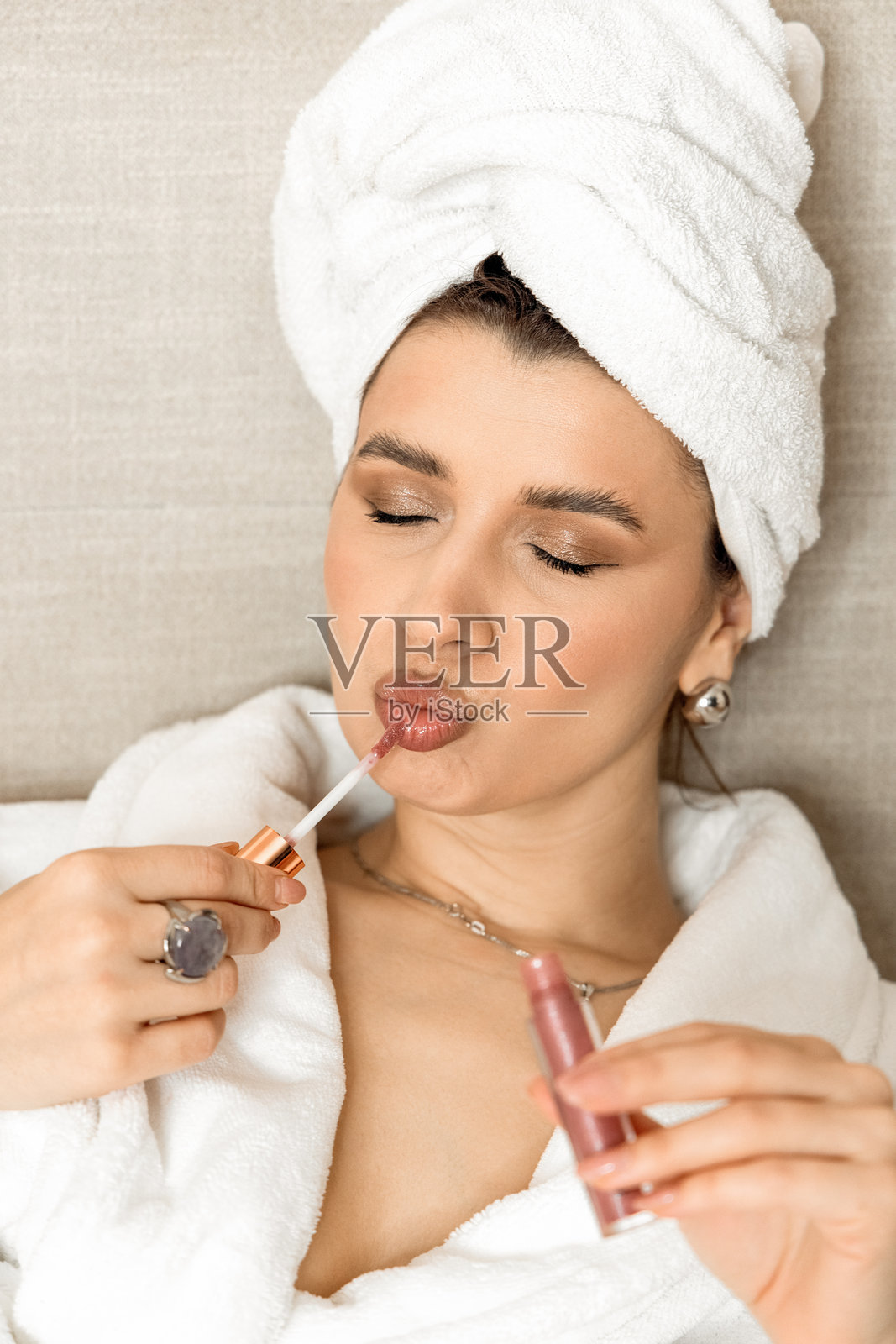 女人在床上醒来。女孩头上裹着毛巾，穿着浴袍，坐着，手里捧着咖啡杯和日记本。女性正在涂唇膏，使用精华液或拿着小闹钟。女人伸展双手，揉眼睛。照片摄影图片