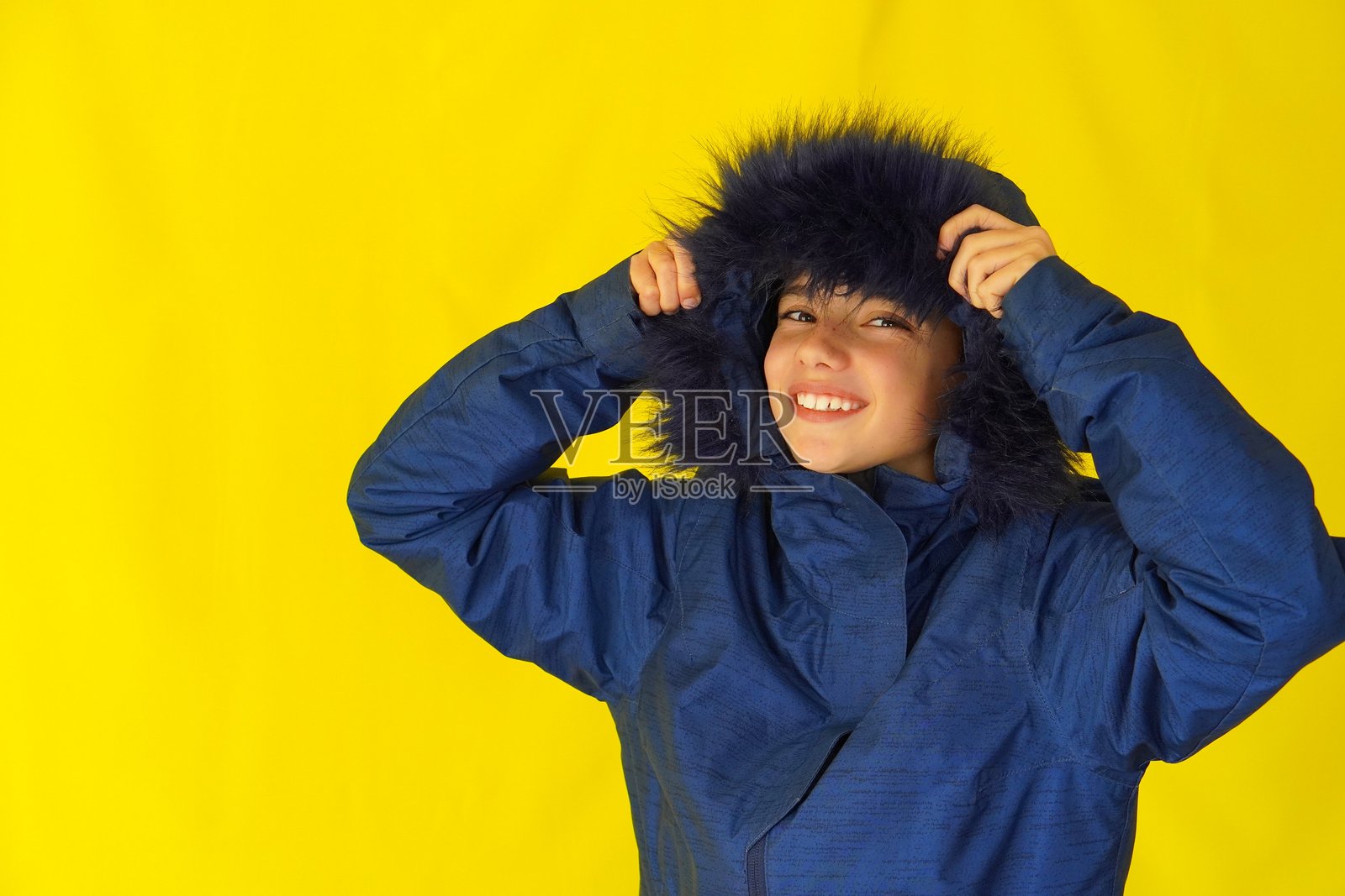 快乐的青春期前男孩，脸上有雀斑，正在为寒冷和下雪的天气穿上蓝色夹克的帽子。照片摄影图片