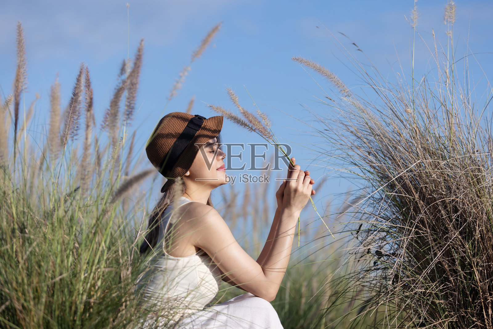 一位穿着白色裙子和帽子的年轻女性坐在高草丛中，手握一根草茎，凝视远方。照片摄影图片