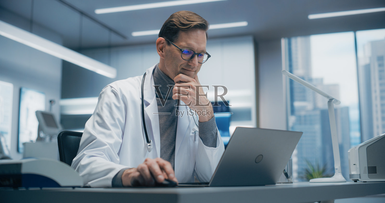 严肃的男性医生穿着白大褂和眼镜，坐在桌子前。脖子上挂着听诊器，专注地查看笔记本电脑屏幕。现代医疗办公室，明亮的灯光。专业精神与先进的医疗保健。照片摄影图片