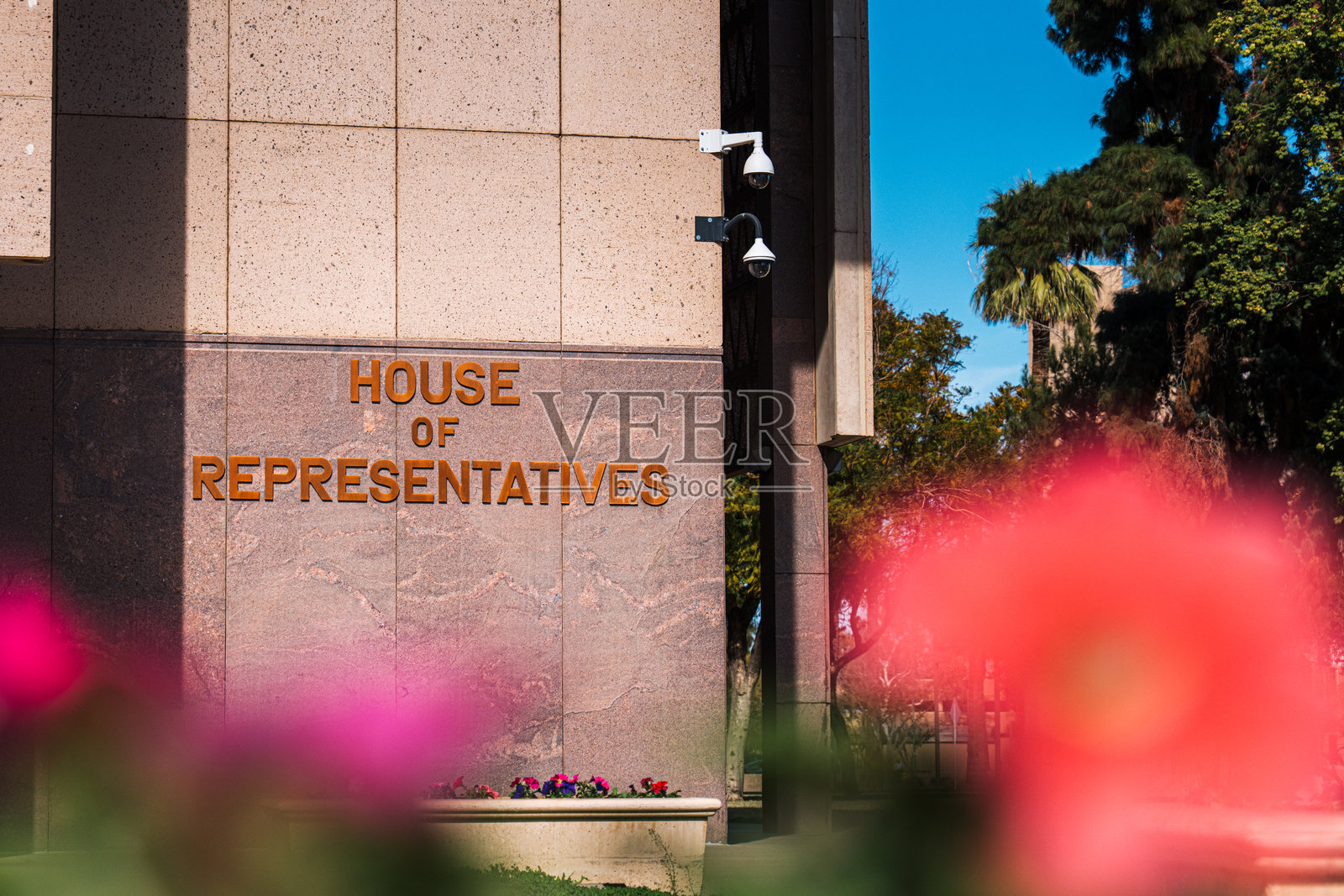阳光明媚的日子里，凤凰城州议会大厦前的鲜艳色彩的矮牵牛花与“众议院”标志和监控摄像头照片摄影图片