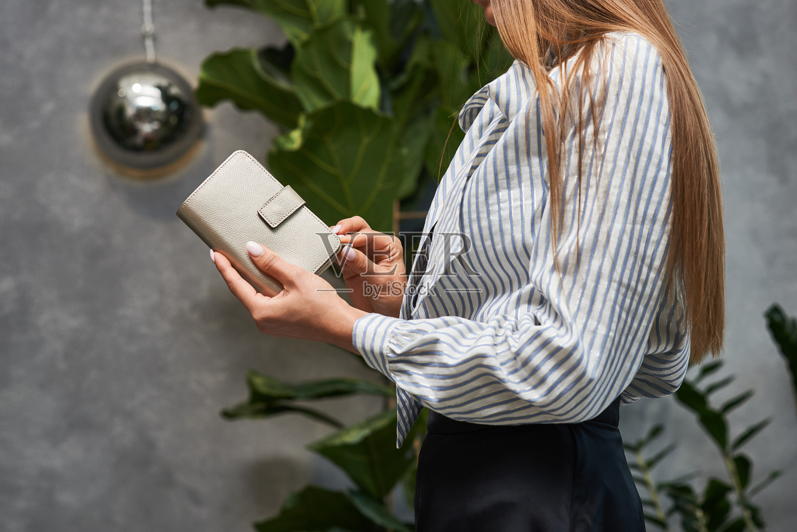 优雅的女性穿着条纹上衣，在室内绿植环境中手持时尚皮革钱包，展现出精致的时尚理念照片摄影图片