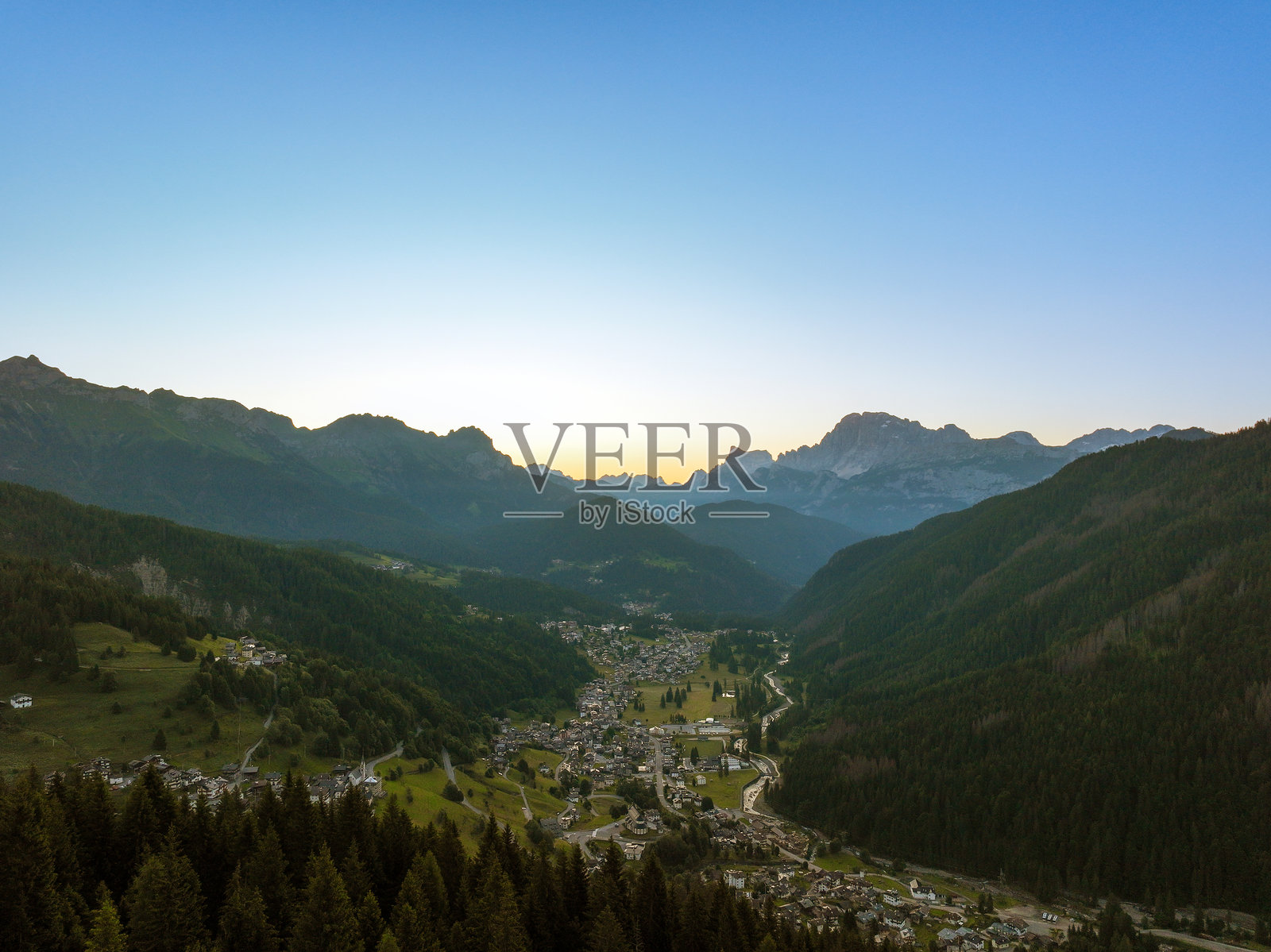 意大利多洛米蒂山脉的夏季天空中明亮的太阳圆盘，空中俯瞰山脚下的度假小镇。山脉的壮丽景观，绿色的针叶林和草地。旅游旅行概念。照片摄影图片