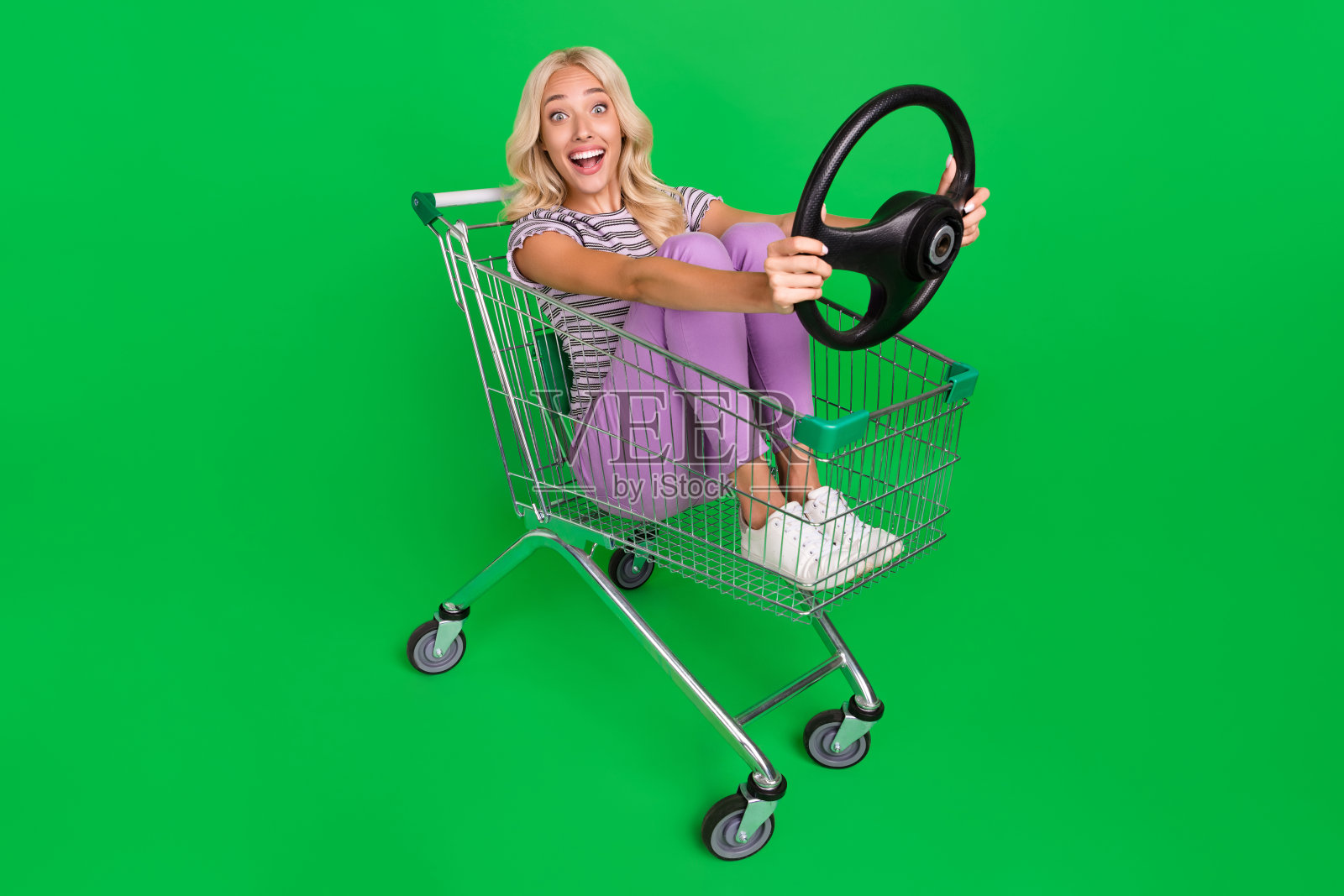 惊讶而积极的人全身照，坐在购物车上，手握车轮，背景为绿色。照片摄影图片