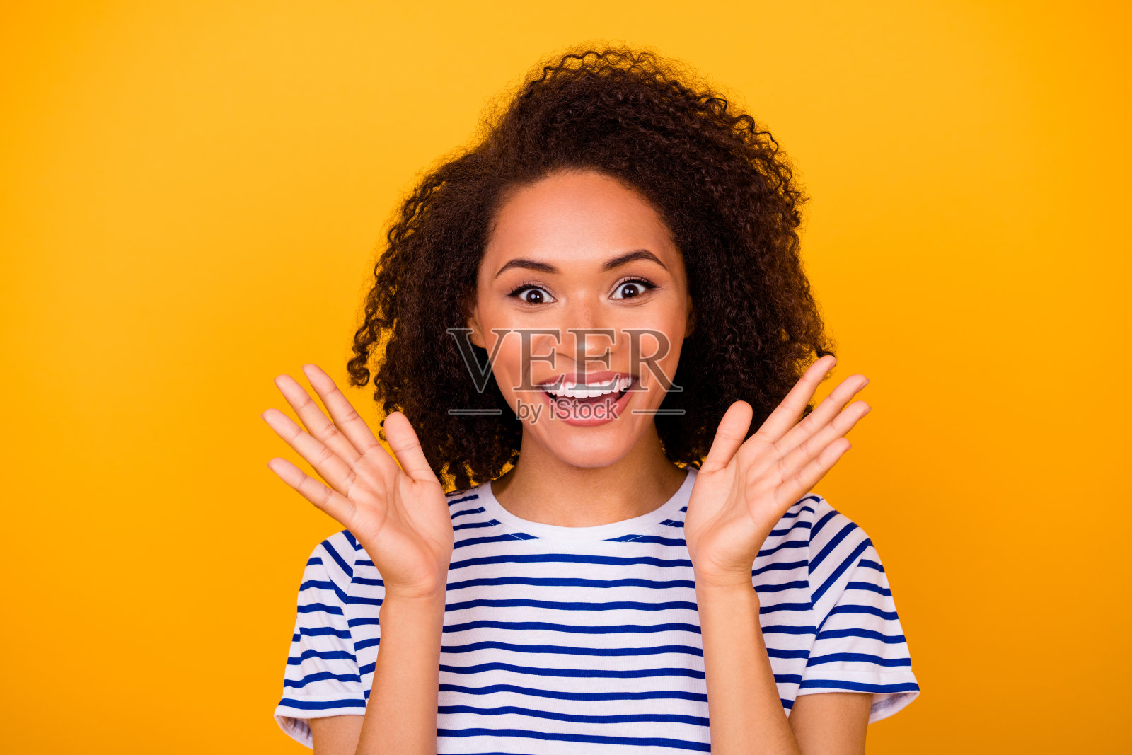 一位有趣的女士穿着白色T恤，手指下巴，微笑着举起双臂，背景为黄色。照片摄影图片