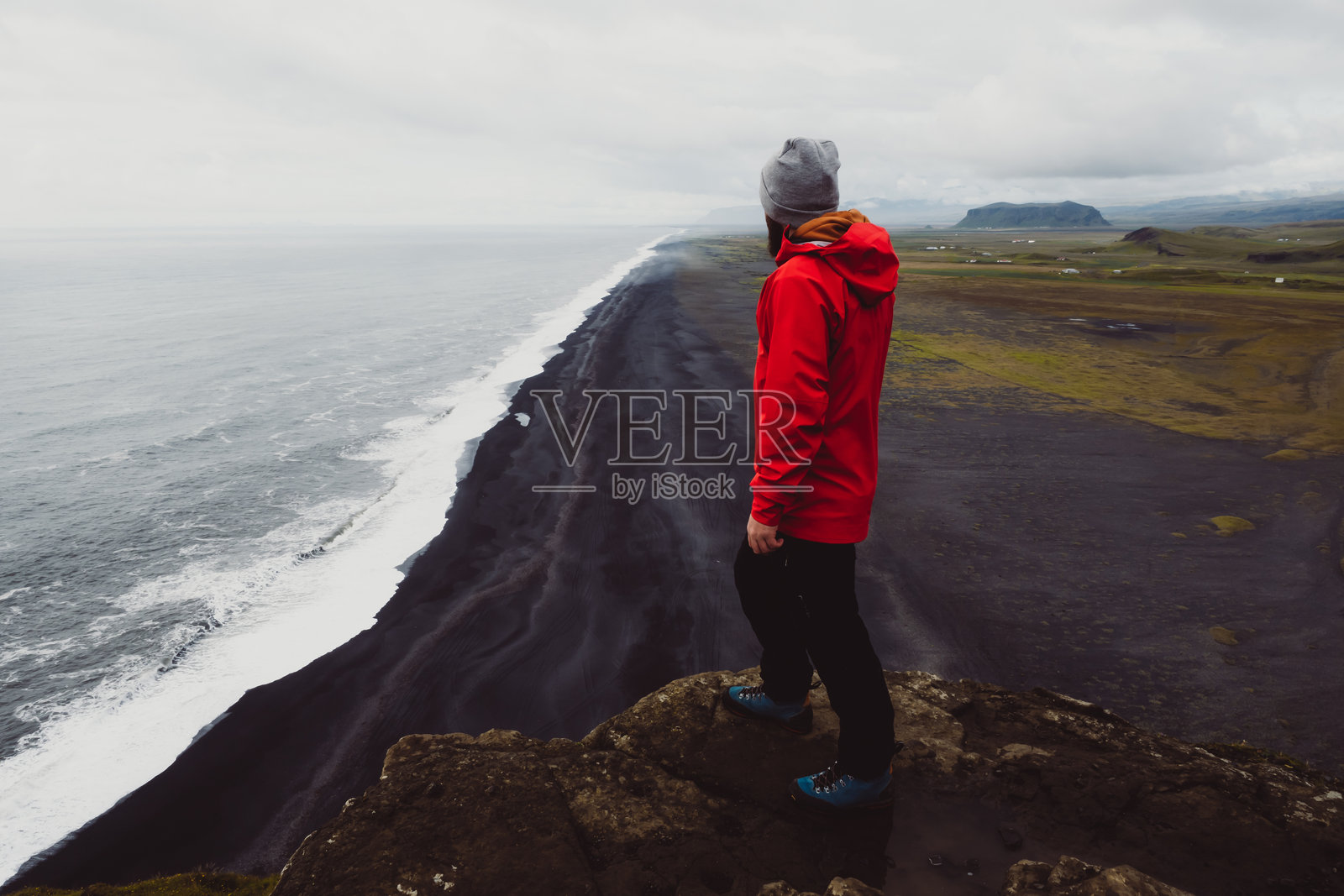 穿着红色夹克的探险者俯瞰冰岛黄金圈内的迪尔霍拉伊灯塔悬崖上雷尼斯菲亚拉黑沙滩的美丽海岸线照片摄影图片