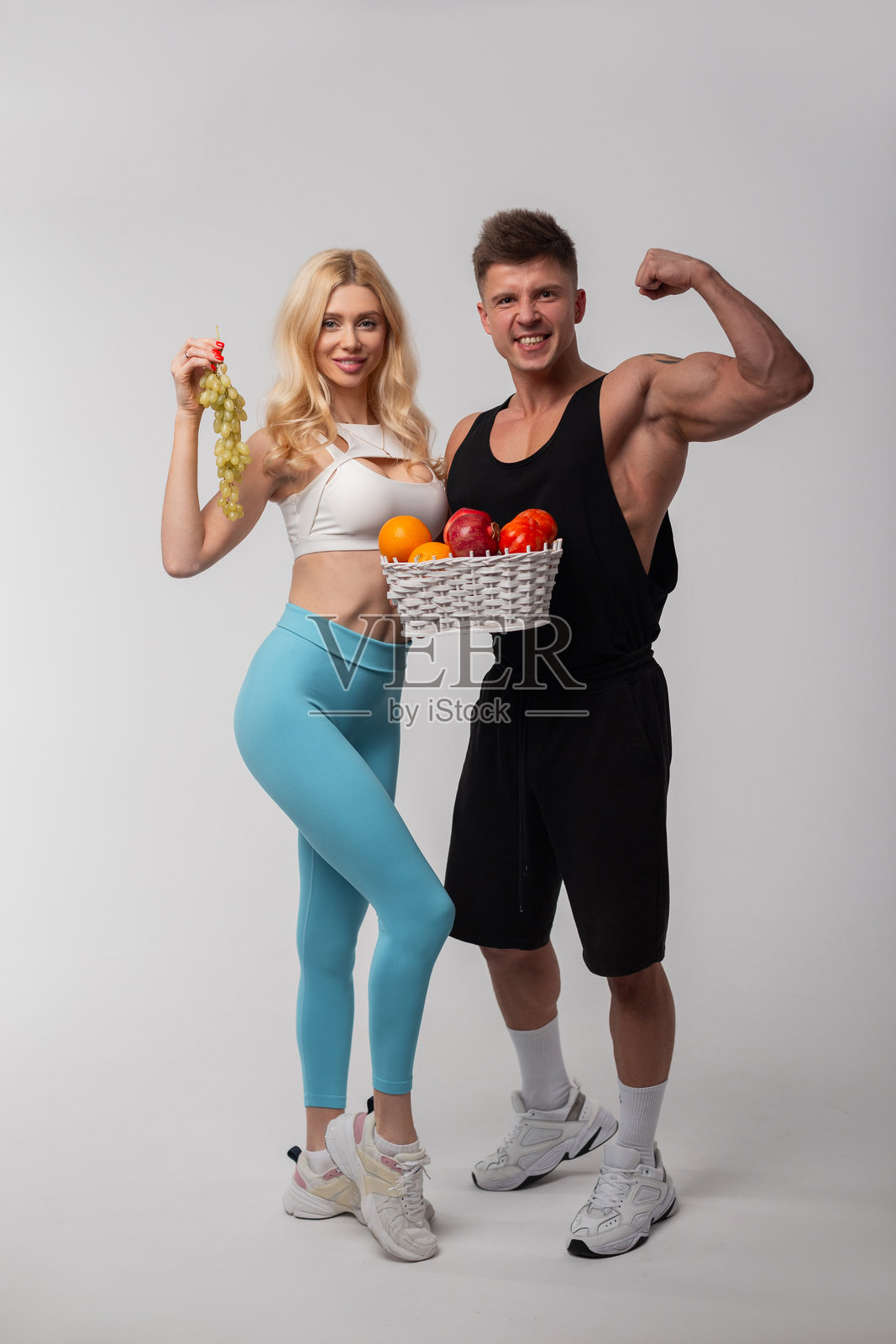 强壮美丽的健康健身情侣，金发女孩和肌肉型男在工作室里摆姿势，手里拿着一篮水果，包括葡萄、苹果和橙子。健康饮食，维生素。照片摄影图片
