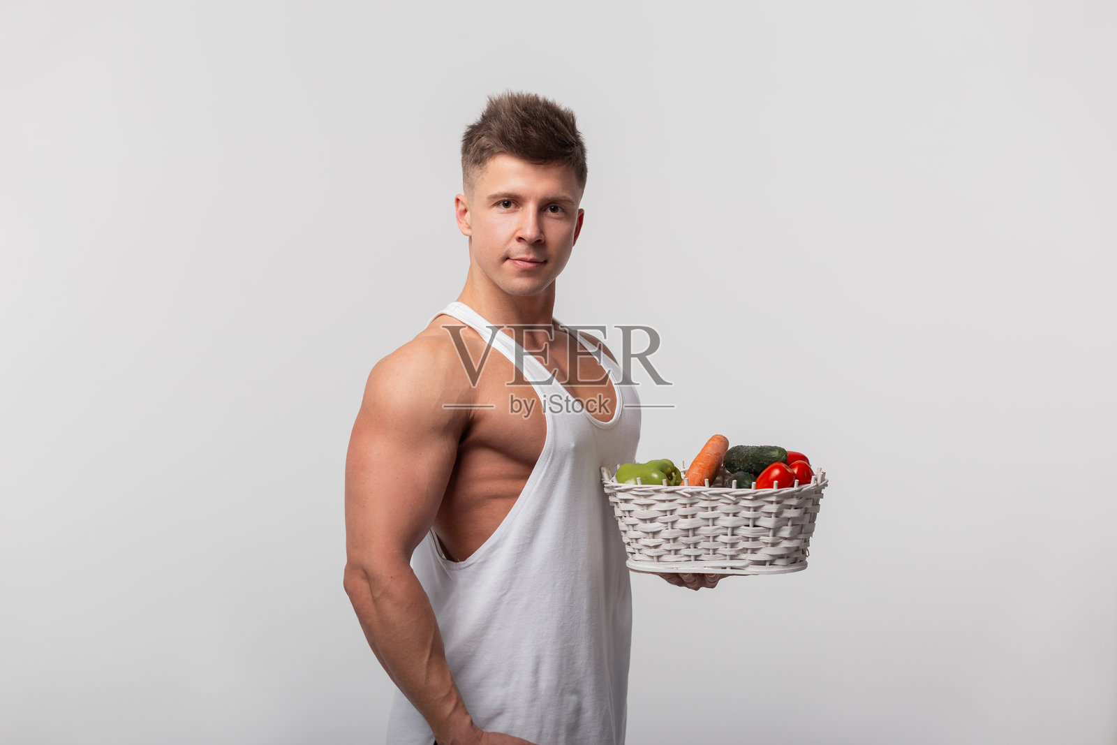 健康英俊强壮的运动男士手持装有蔬菜的篮子，背景为灰色。生食饮食和健康生活方式。天然维生素。照片摄影图片