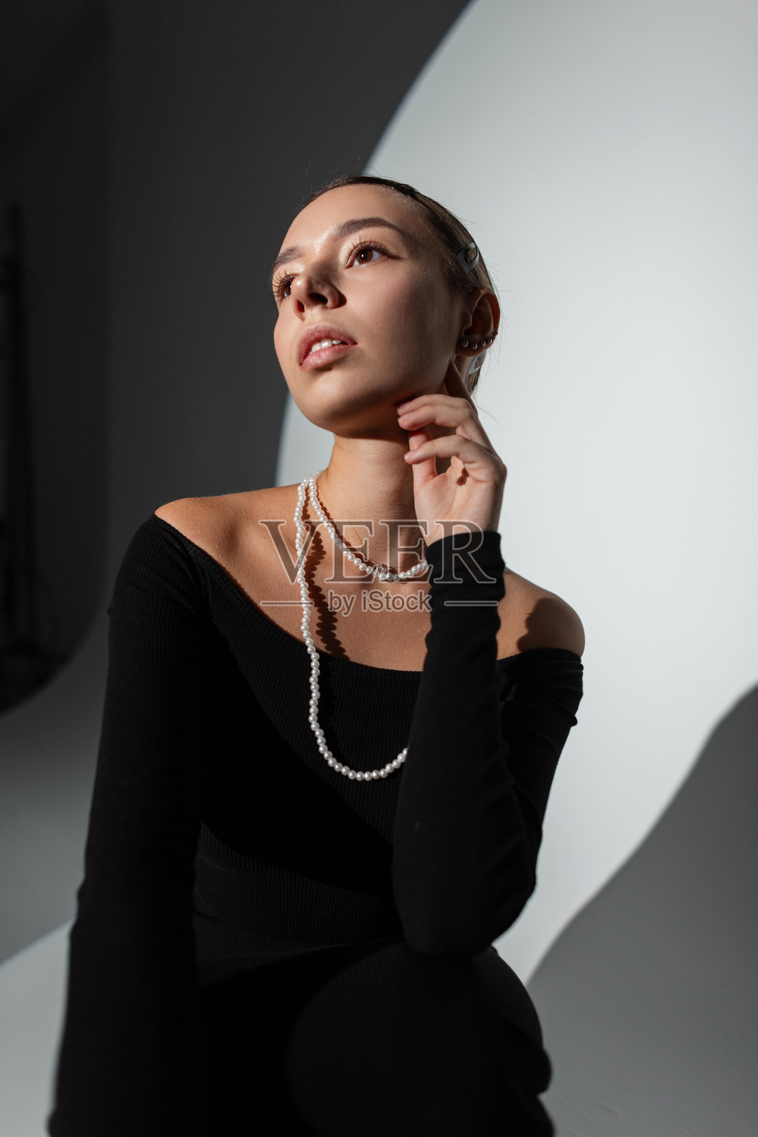 优雅美丽的年轻女性穿着时尚的黑色连衣裙，佩戴项链，坐在暗背景的工作室中，摆姿势，周围有圆形硬光。迷人的美丽女士。照片摄影图片