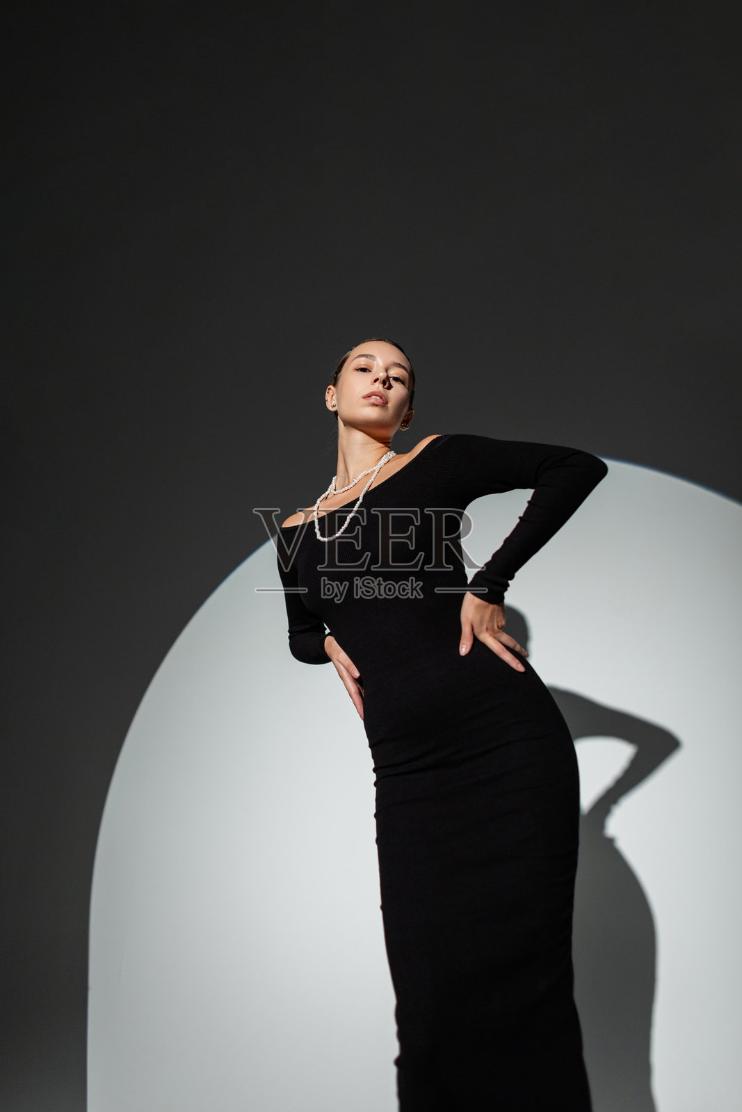身穿时尚长款黑色连衣裙的美丽年轻女性在暗色背景的工作室中摆姿势，周围有圆形硬光照明。漂亮女孩。照片摄影图片
