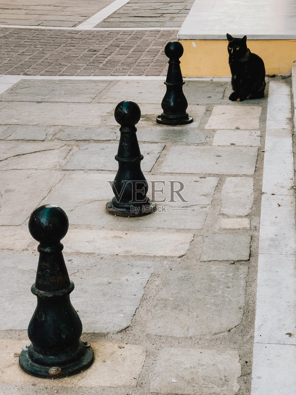 黑猫安静地躺在石铺街道的路障旁，享受阳光，沉浸在城市景观中，享受户外宁静的时刻，感受着宁静的城市氛围。黑猫坐着。照片摄影图片