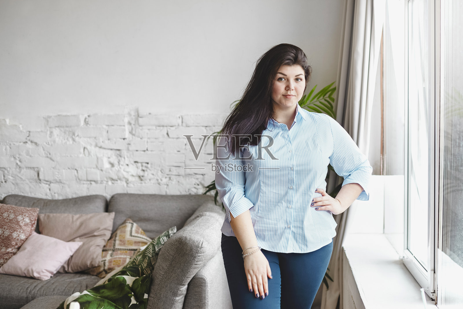 二十多岁的美丽丰满的棕发白人女孩，穿着 XXL 蓝色条纹衬衫和紧身牛仔裤，站在家中大窗户旁，背景是砖墙和沙发。照片摄影图片
