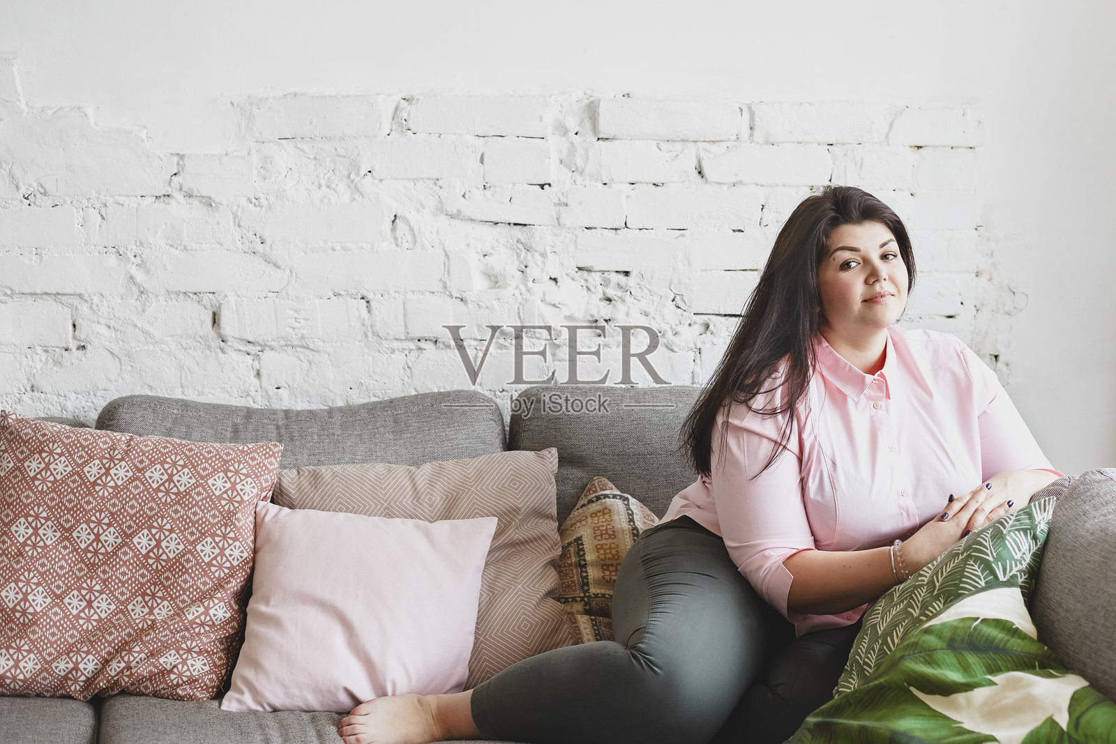 快乐美丽的年轻胖女孩，留着松散的黑发，穿着宽大的粉色衬衫和紧身牛仔裤，舒适地坐在沙发上，双手交叉，留有文本空白。照片摄影图片