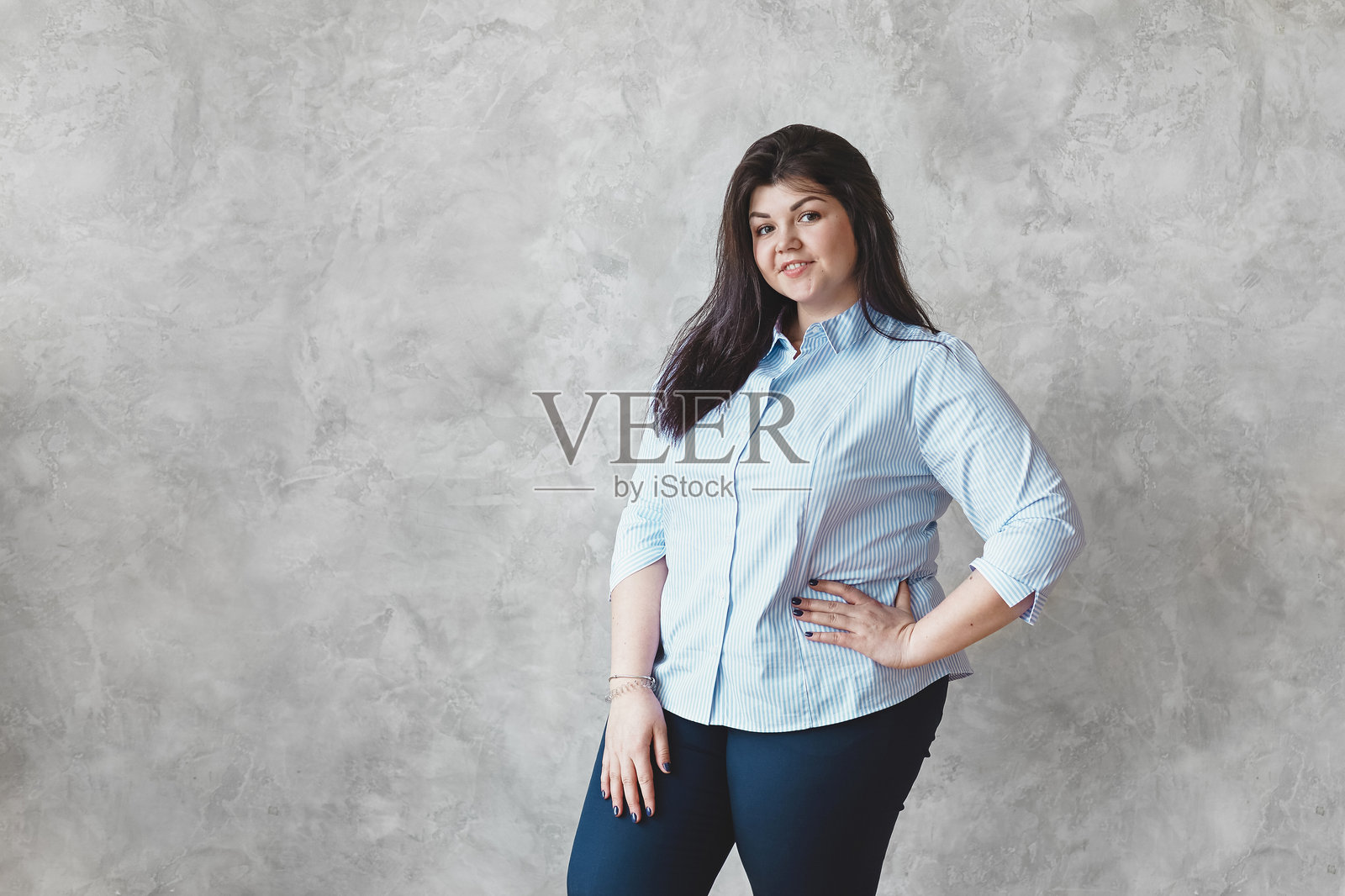 美丽的年轻丰满女性，长发披肩，站在空白的纹理墙背景前，留有空白空间供您使用。身体积极性概念。照片摄影图片