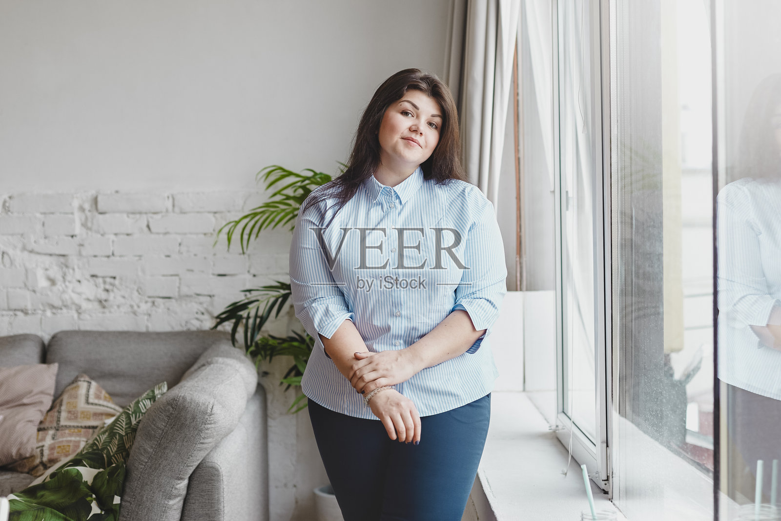 人们、超重、风格和时尚概念。室内拍摄一位迷人的年轻超重棕发女性，穿着大号蓝色衬衫和牛仔裤，站在窗边的客厅内。照片摄影图片