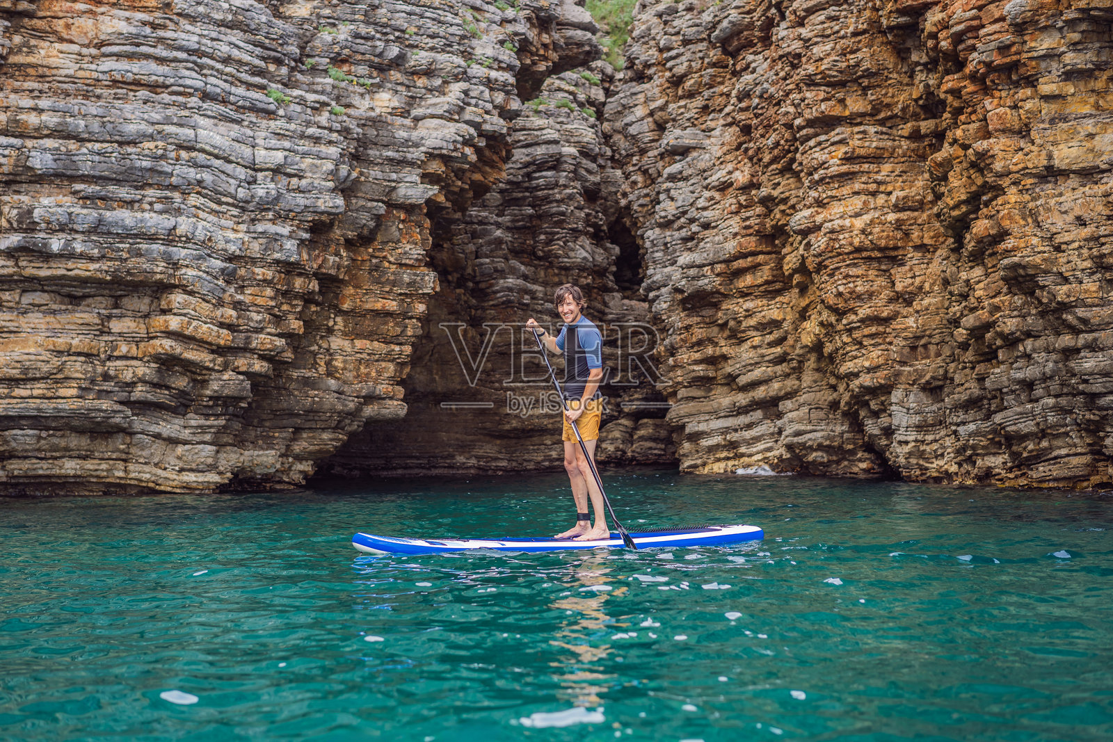 年轻人在黑山的蓝色海水中，岩石间享受立式划水的乐趣。照片摄影图片