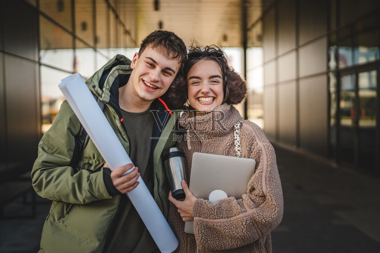 一对情侣在户外拥抱着走，手里拿着笔记本电脑和保温瓶。照片摄影图片