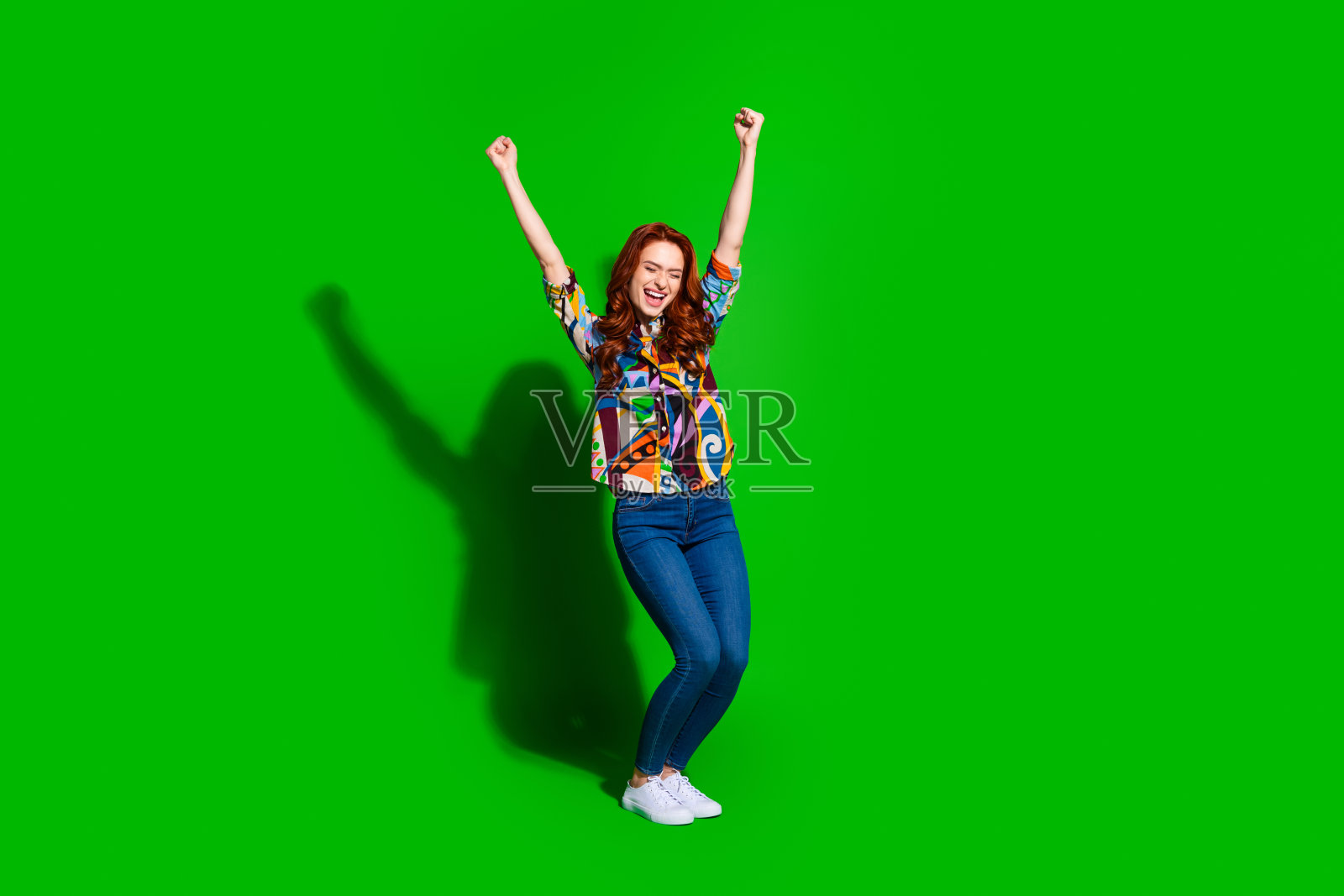 微笑的年轻女性在充满活力的绿色背景下，双臂高举，穿着色彩斑斓的上衣，庆祝快乐的时刻照片摄影图片
