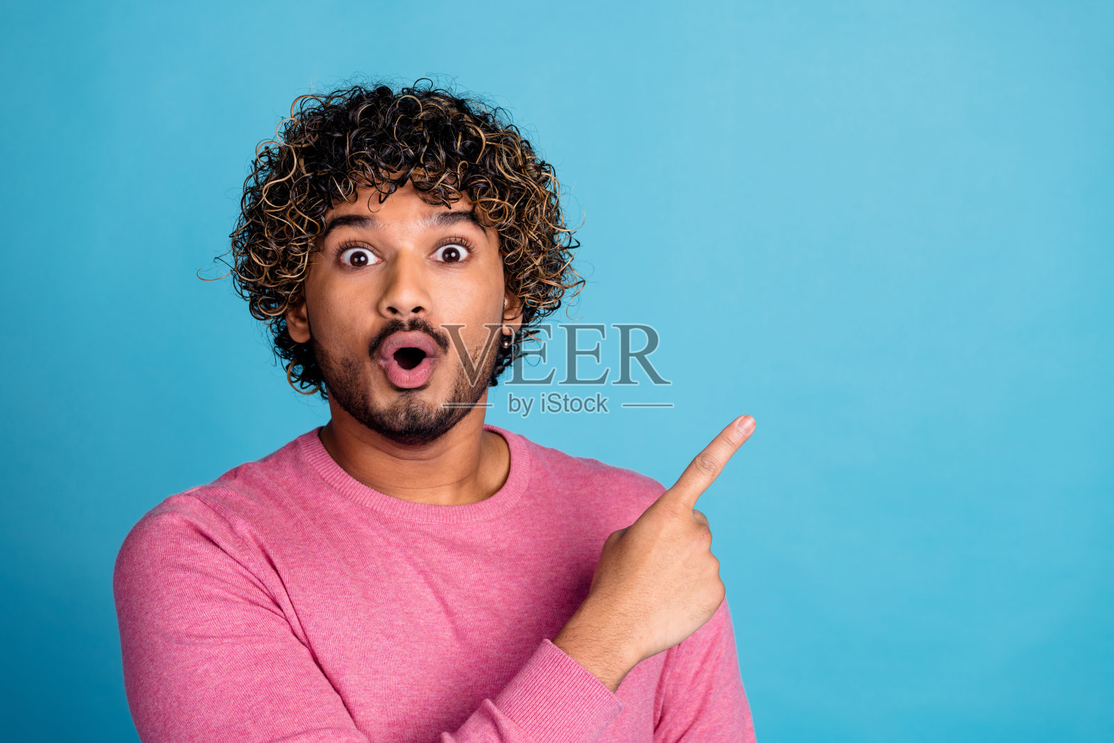 时尚的年轻男子，卷发，穿着粉色毛衣，惊讶地用食指指向鲜艳的蓝色背景。照片摄影图片